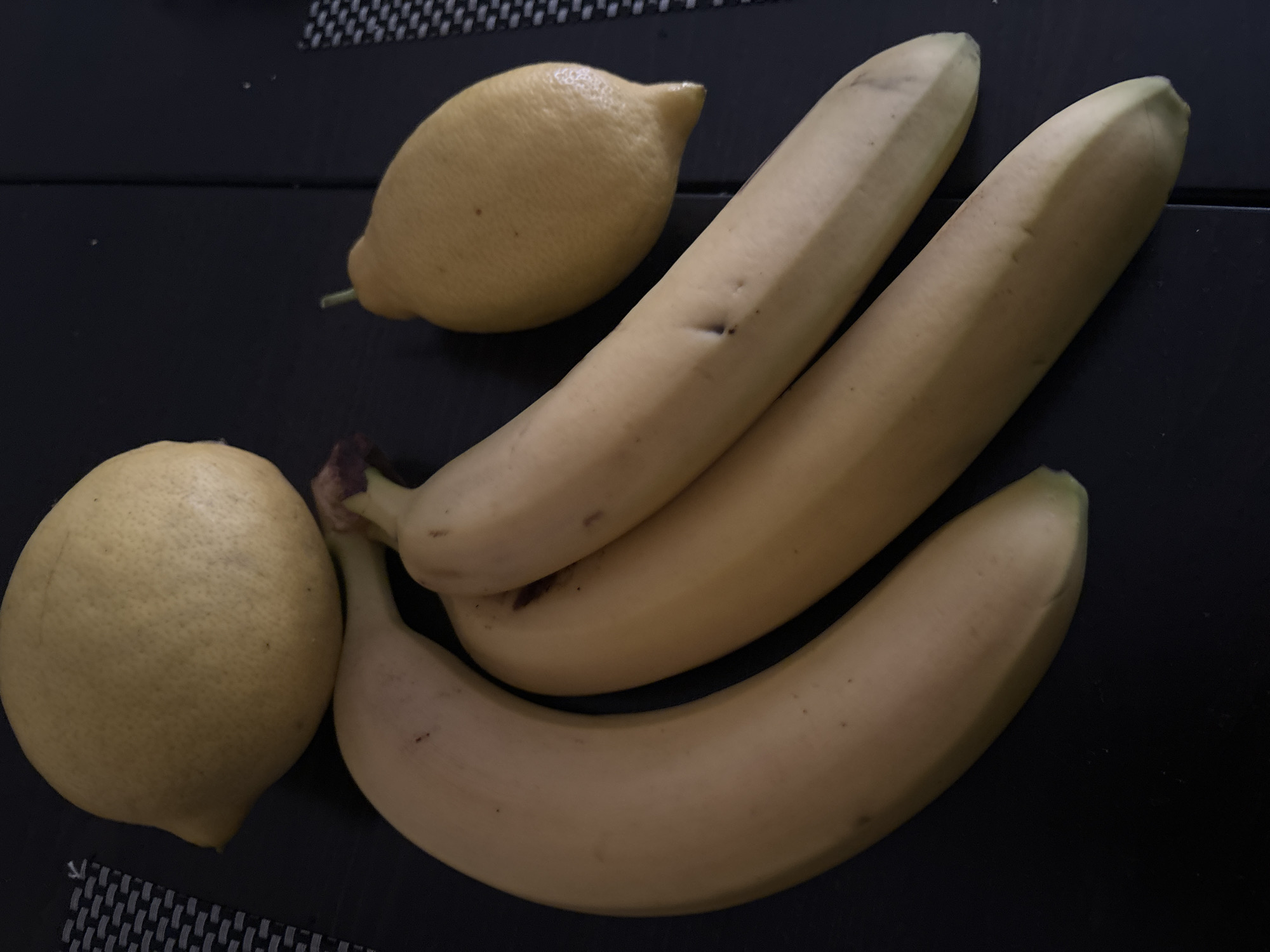 The image presents a still life composition featuring two lemons and three bananas arranged on a dark, flat surface. This surface appears to be a table or countertop, with a subtle dark grey and black checkered pattern visible in the upper and lower left corners, suggesting a placemat or integrated design.
In the upper left portion of the frame, one lemon is visible, characterized by its bright yellow color and slightly elongated shape. Below and to its left, a second, larger lemon is positioned, displaying a slightly lighter, more muted yellow color and a distinctively textured skin.
To the right of the lemons, a cluster of three ripe bananas lies horizontally across the frame. The bananas are a pale yellow, with a few small brown spots indicative of their ripeness. They are partially overlapping, with one banana positioned slightly above the other two. The dark, brownish-purple stems of the bananas are visible at their point of connection.
No activity or event is taking place; the fruits are static. The image provides no visual cues regarding the time of day, weather conditions, or any emotions. No text is visible within the photograph. While the location context of Budva, Montenegro, is provided, the image itself does not contain any specific visual elements to confirm this geographic setting.