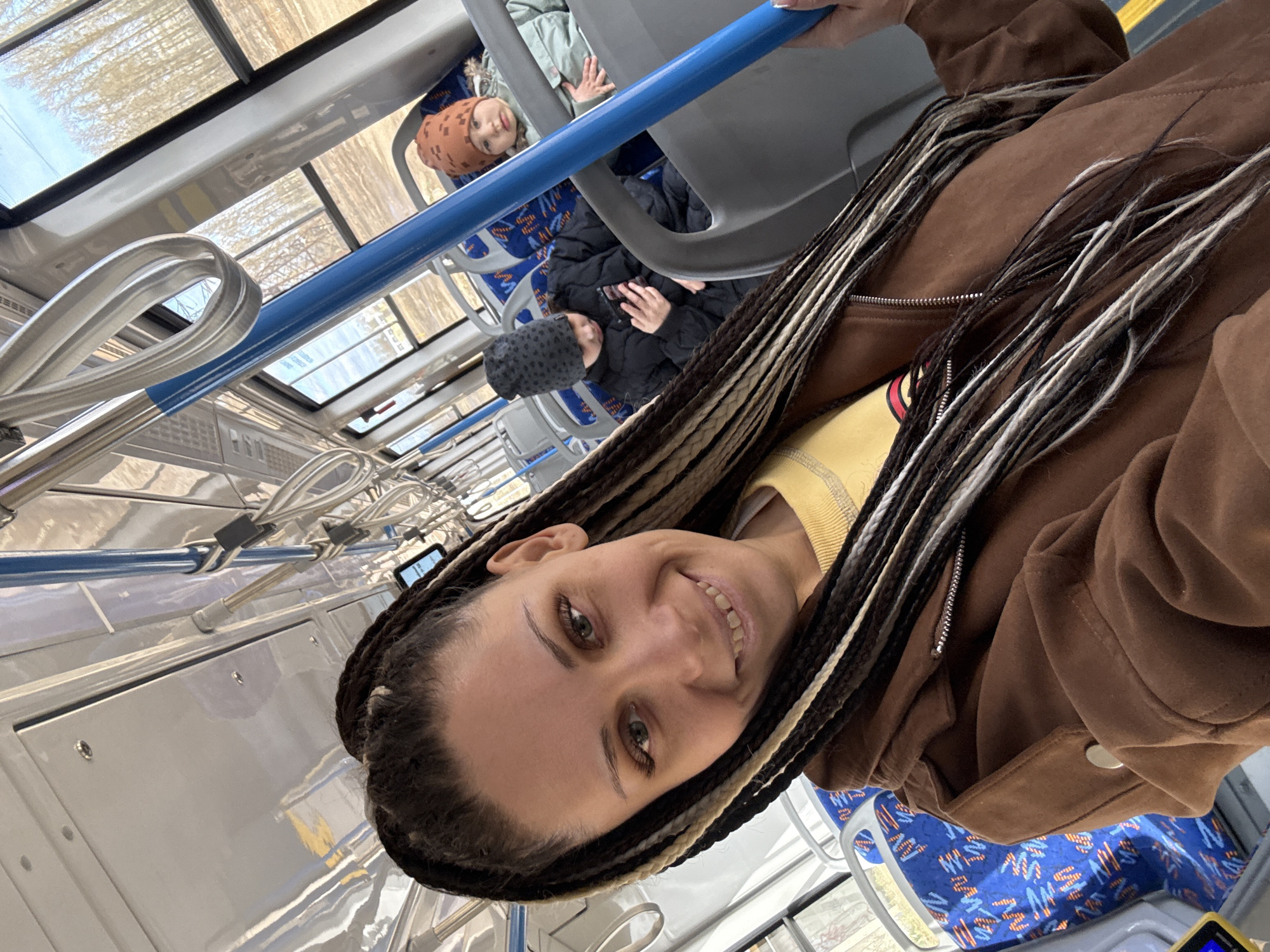 A woman with long, multi-toned braids is posing for a selfie inside a public transportation vehicle. She is wearing a brown jacket and a yellow shirt. Behind her, other passengers are seated. The vehicle has a blue handrail and overhead windows. The seat covers are blue with an orange pattern. The location is Rusanovo, Russia, but this is not verifiable from the image.