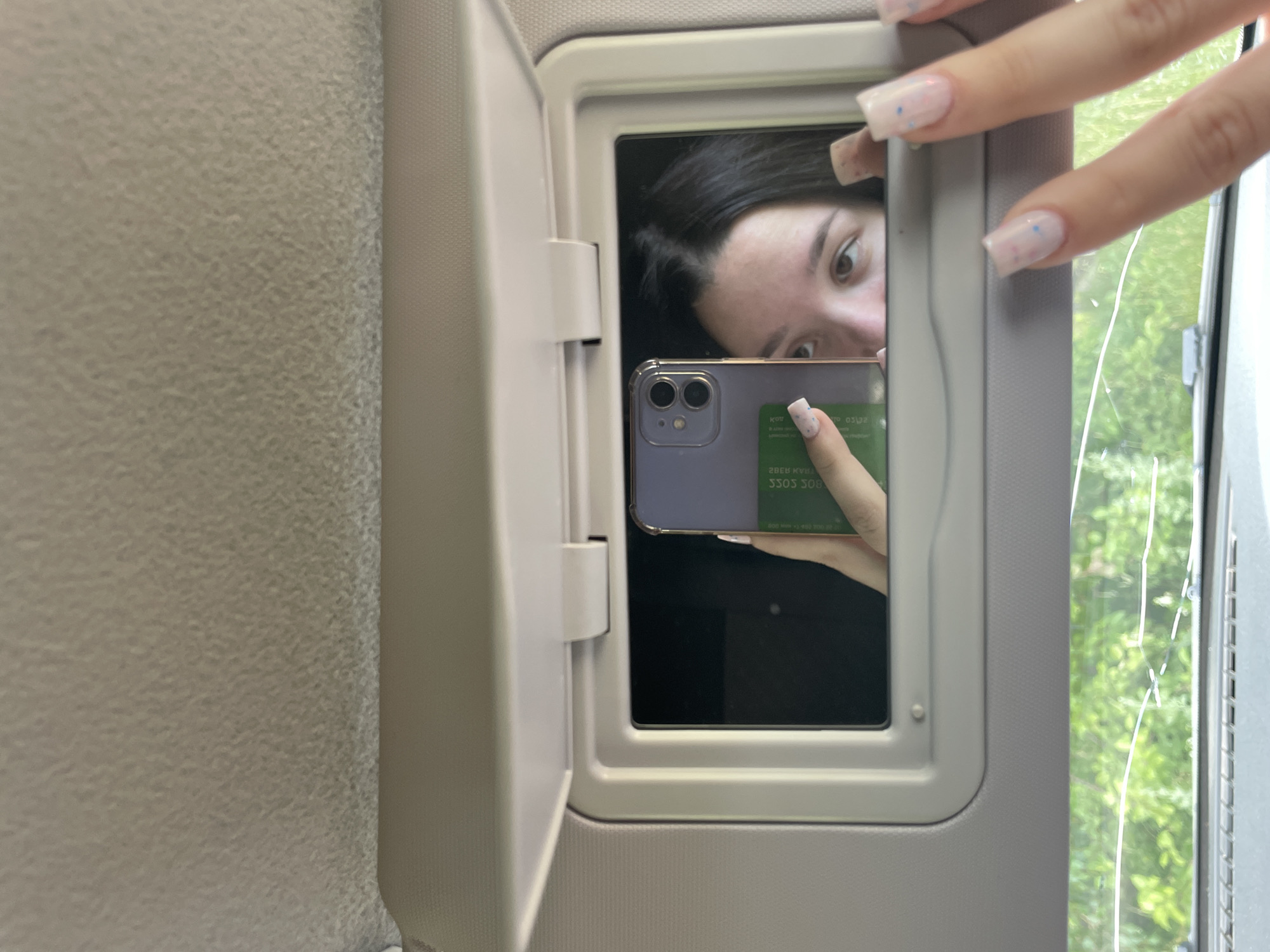 A young woman is taking a selfie in the visor mirror of a vehicle. She has long, dark hair and is looking at the camera with a curious expression. She is wearing a light-colored top. Her right hand, with light-colored, square-shaped nails adorned with small blue speckles, is holding a purple smartphone. The phone screen is visible in the mirror, showing the front-facing camera's view of the woman and her phone. A green card with white text is attached to the back of the phone.
The background of the mirror reflects trees and greenery, suggesting an outdoor setting, possibly a road trip. The visor is open, revealing the interior of the vehicle, which appears to be a car or van, with a textured gray fabric lining. The overall lighting suggests it is daytime, with natural light streaming in from a window to the right of the frame.
The visible text on the green card reads "28E8 KVA", "3303 308", and what appears to be "OOS O3". The text is reversed due to being viewed in a mirror. The image is taken from a slightly high angle, looking down towards the visor. The mood is casual and self-reflective, common for a personal selfie.