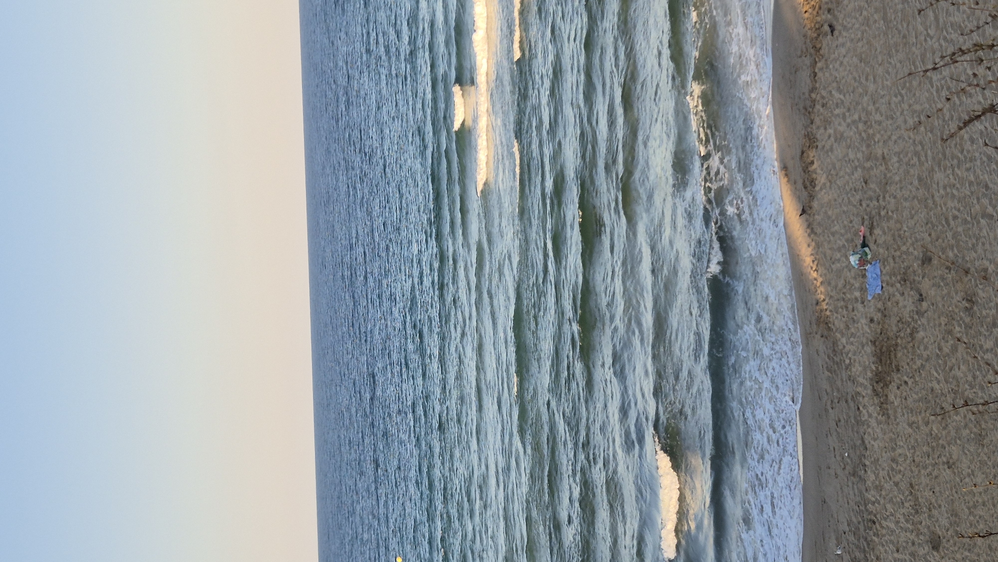 A serene beach scene in Primorsko, Bulgaria, captures the vastness of the sea under a clear sky. The ocean, a deep blue, is gently rippled by incoming waves, creating a rhythmic pattern that stretches to the horizon. The beach, composed of fine sand, slopes towards the water, with the gentle tide washing ashore. In the foreground, a solitary figure lies on a light blue towel, seemingly at rest, surrounded by their belongings. A few birds peck at the sand near the water's edge, adding a touch of wildlife to the tranquil setting. The sun casts long shadows across the sand, indicating it may be late afternoon or early morning.