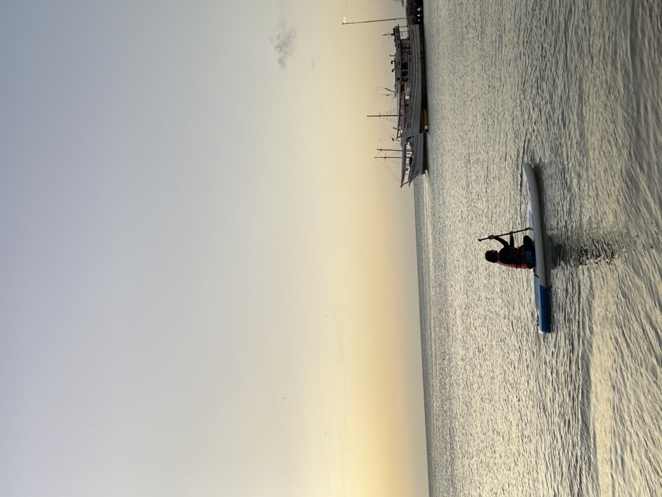 A lone paddleboarder glides across the shimmering water, paddle in hand, towards a large, docked ship with visible masts. The sky transitions from a pale blue to a warm yellow as the sun likely sets or rises. The water reflects the sky's hues, creating a peaceful, expansive scene. The boat appears as a traditional vessel, contrasting with the modern paddleboard.