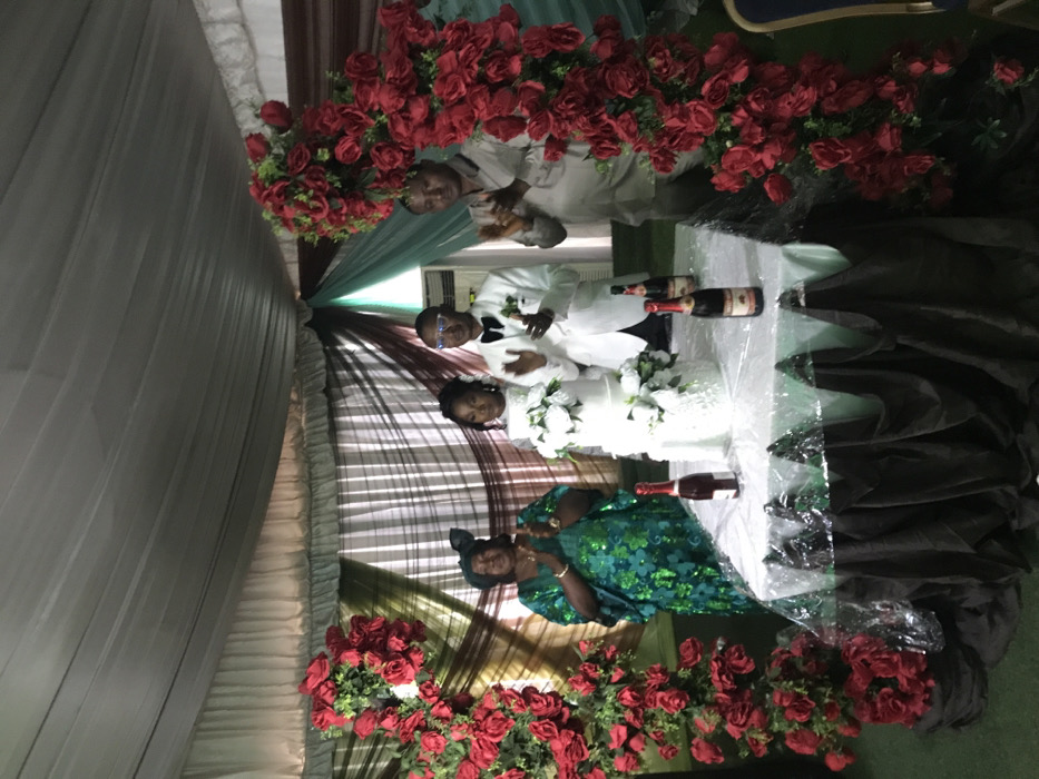 This image captures a lively indoor celebration, likely a wedding reception, featuring five individuals gathered around a decorated table. The overall scene is festive, adorned with abundant red roses and elegant drapery.
In the center of the frame are what appear to be the bride and groom. The groom, a man wearing a crisp white suit, a white shirt, and a black bow tie, stands behind a white-covered table. He is wearing glasses and looking towards the viewer, holding something indistinct in his right hand. Beside him, the bride, in a white dress adorned with white floral accents, also stands behind the table, her gaze directed towards the camera. A multi-tiered white wedding cake, decorated with white flowers, is prominently displayed on the table directly in front of the couple.
To the left of the central couple, an older woman is visible, dressed in a vibrant green and blue patterned garment. She is looking towards the couple. Further to the left, at the bottom corner, only the head and shoulders of another person with a dark head covering are partially visible. On the table, various bottles, likely sparkling wine or champagne, are placed; two are situated behind the bride and groom, and one is to the left of the cake. The label on one of these bottles partially reveals the word "CHANDON."
To the right of the central couple, an older man is partially obscured by decorative elements. He is wearing a light-colored, possibly beige or cream, traditional outfit and appears to be holding a glass.
The background of the scene is tastefully decorated with draped fabrics in shades of white, dark brown or maroon, and teal green, providing a soft and elegant backdrop. An air conditioning unit is visible on the wall behind the groom. Framing the celebratory area on both the left and right sides are extensive arrangements of bright red artificial roses and lush green foliage, adding a vibrant touch to the decor. The lighting suggests an indoor setting or an evening event, with artificial light illuminating the scene.