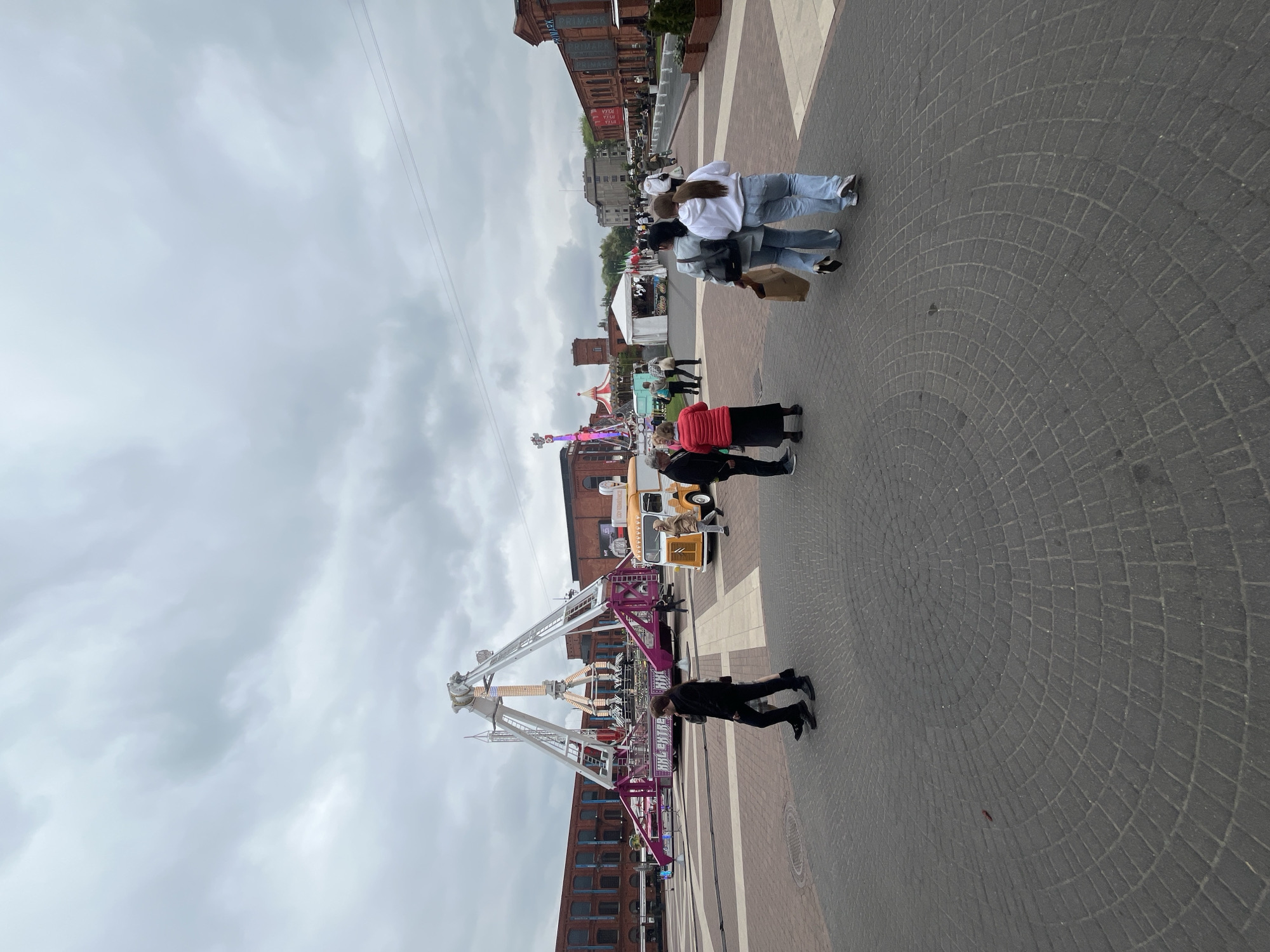 The image depicts a scene at Manufaktura, a shopping and entertainment complex in Łódź, Poland.
The foreground shows a pedestrian area paved with gray cobblestones. Several people are walking, including a group of three wearing jeans and light-colored jackets, and a person in a black outfit standing near a fairground ride. Another group consists of a person in red and one in black near a yellow-painted vehicle.
A fairground ride with white metal supports and a rotating element with lights is positioned in the center of the image.
In the background, several red brick buildings are visible, including one labeled "PRIMARK." Additional tents and figures suggest other fairground attractions and more people present in the distance. The sky is overcast.