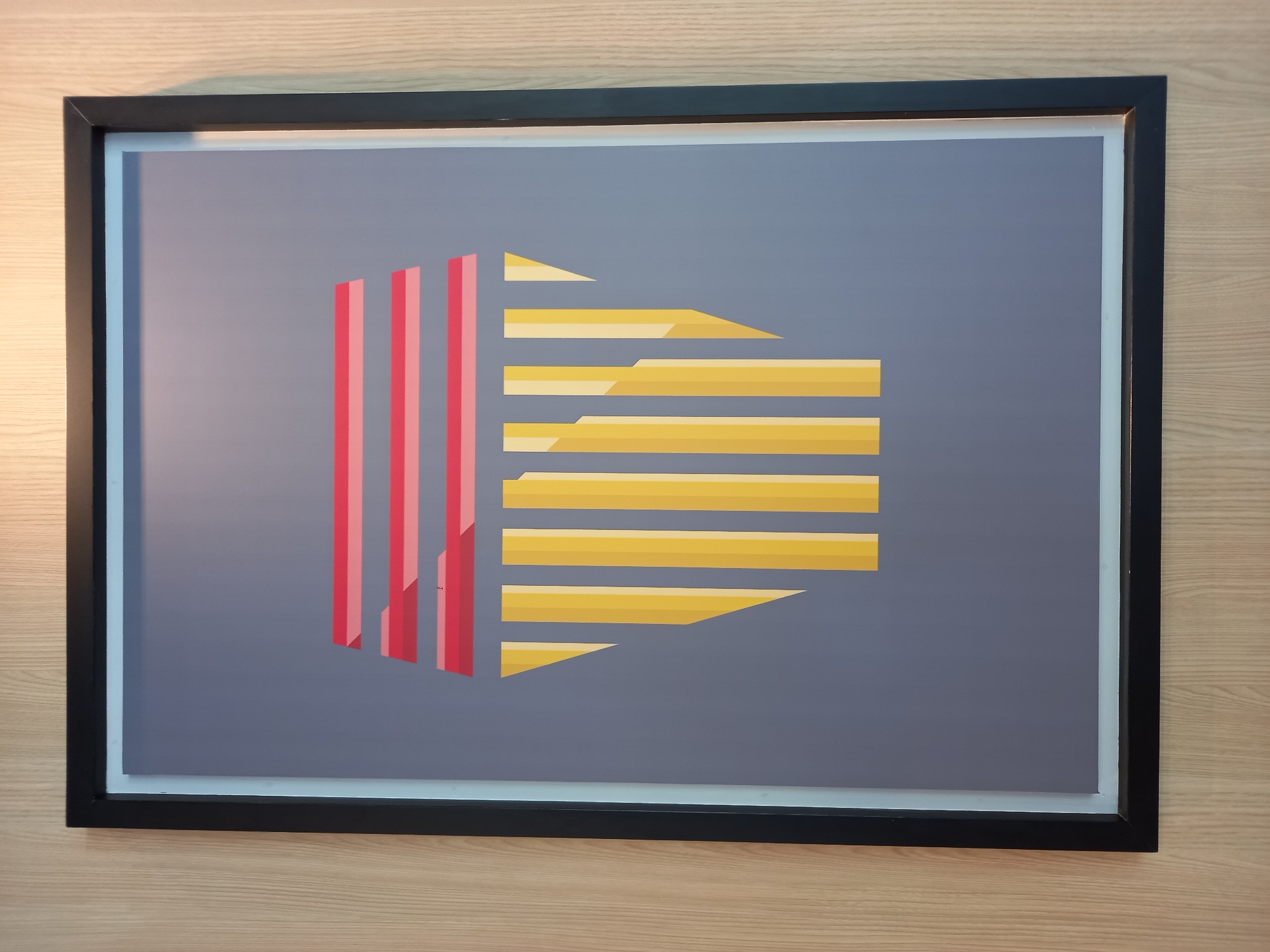 A framed modern art print is hanging on a light wood-paneled wall in Baan Bo Phut, Thailand. The print has a gray background with a geometric design in red, pink, and yellow. The design resembles a stylized column or facade with horizontal red and pink stripes at the top and vertical yellow stripes below. The artwork is centered within the frame, which is black with a thin light blue matting. The lighting in the scene is soft, suggesting an indoor environment. There are no people or animals visible in the image.
