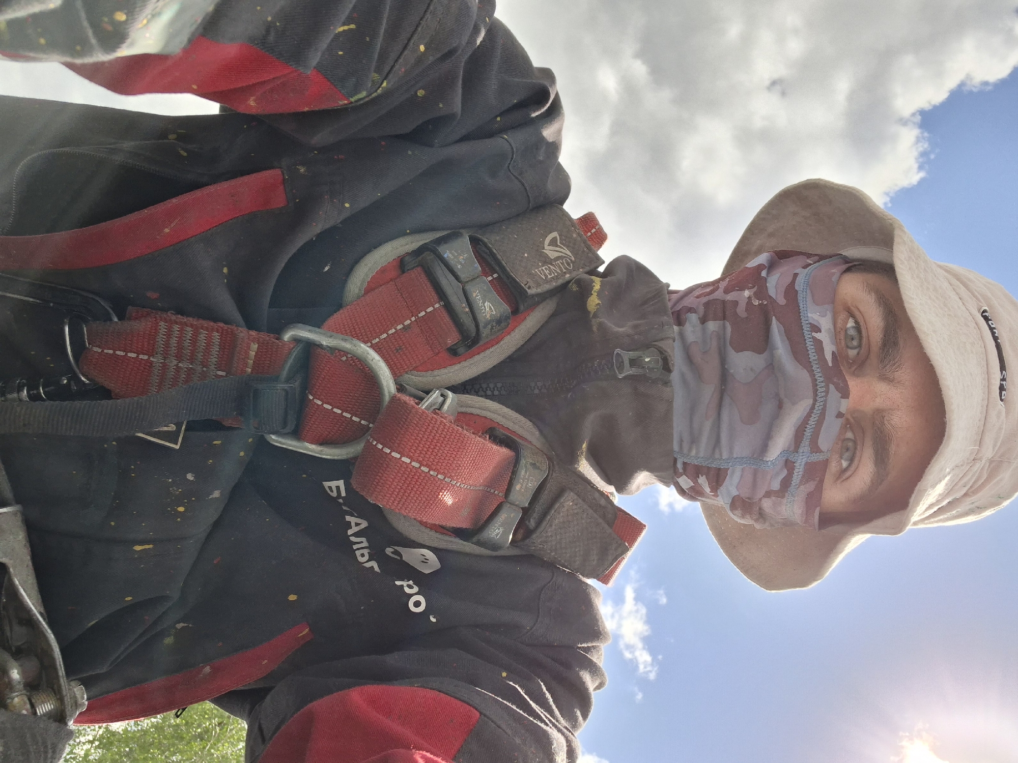 A low-angle, self-portrait shows a person wearing a beige bucket hat and a camouflage-patterned face covering. The person is also wearing a black and red jacket with a red safety harness. Part of the jacket has the lettering "Б.Альб. ро." written on it. The sky is visible in the background with some clouds and the sun is shining. There is some green foliage visible in the lower left corner. The location is possibly the city of Zhabinka, Belarus.
