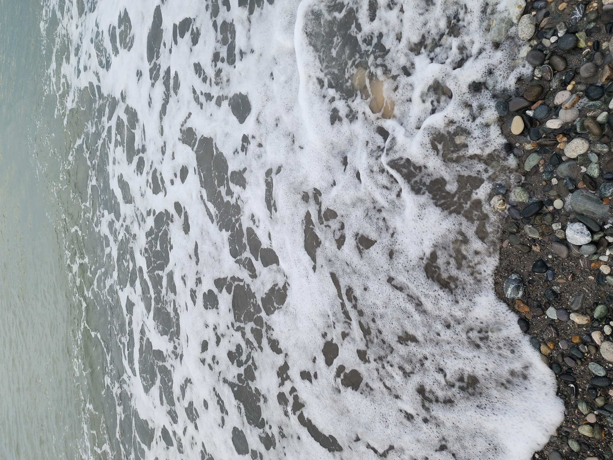 This high-angle, close-up image captures the interaction between the sea and a pebbled beach in Sochi, Russia. The scene is dominated by a frothy layer of white seafoam, likely from a receding or incoming wave, spreading across the wet shoreline. To the left, the calm, grey-green water of the sea is visible, displaying subtle variations in tone.
The right side of the frame features the pebbled beach, composed of numerous rounded stones of various sizes and colors, including shades of grey, black, brown, and white. These pebbles are interspersed with darker sand or earth. The foamy water creates intricate, lace-like patterns as it merges with the wet parts of the shore, contrasting with the drier sections further up the beach.
The image shows no human subjects or man-made objects. The activity depicted is the natural ebb and flow of the tide, with the seafoam marking the water's edge. The lighting appears soft and diffused, suggesting daytime, possibly under an overcast sky, as there are no strong shadows. No text is visible in the photograph. The overall impression is one of natural coastal beauty and the serene, continuous movement of the ocean.