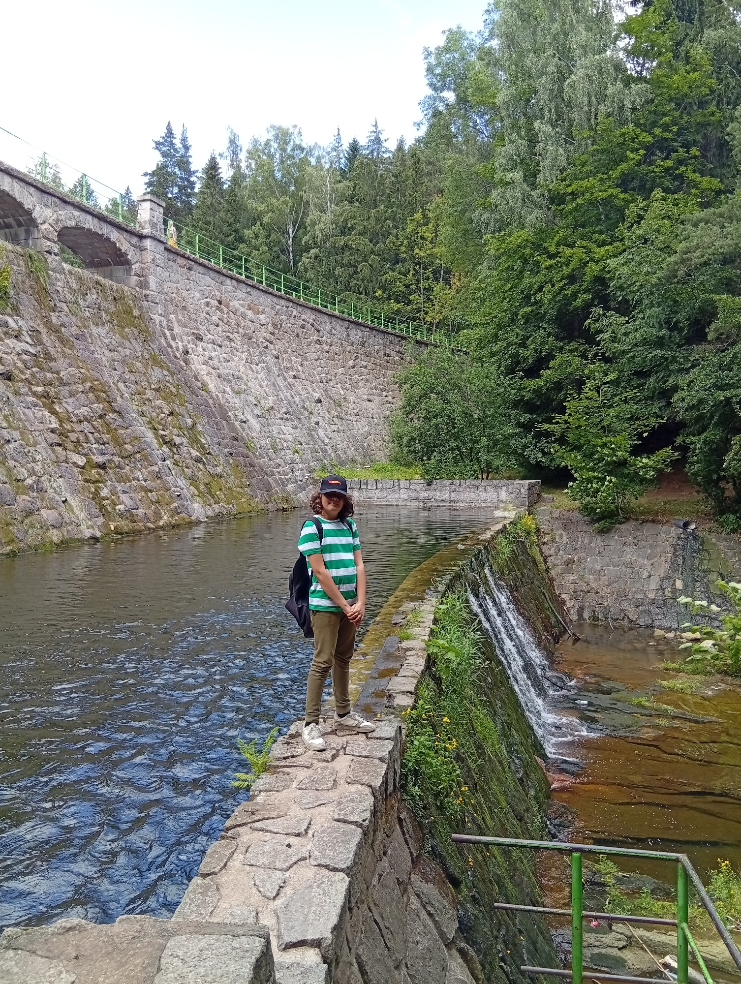 In Karpacz, Poland, a person stands on a stone pathway next to a body of water. The person is wearing a green and white striped t-shirt, olive green pants, white sneakers, and a black baseball cap. They have a black backpack on. The pathway is made of large, irregular stones and runs alongside the water, which appears to be a river or canal. To the right of the pathway, a small waterfall cascades down a stone wall covered in moss and vegetation. Behind the person and the water is a large, imposing stone wall that slopes upwards, constructed of irregular stones. This wall is part of a dam or embankment and has arched openings visible at the top. Lush green trees and foliage cover the landscape behind the stone wall. The sky is overcast.
