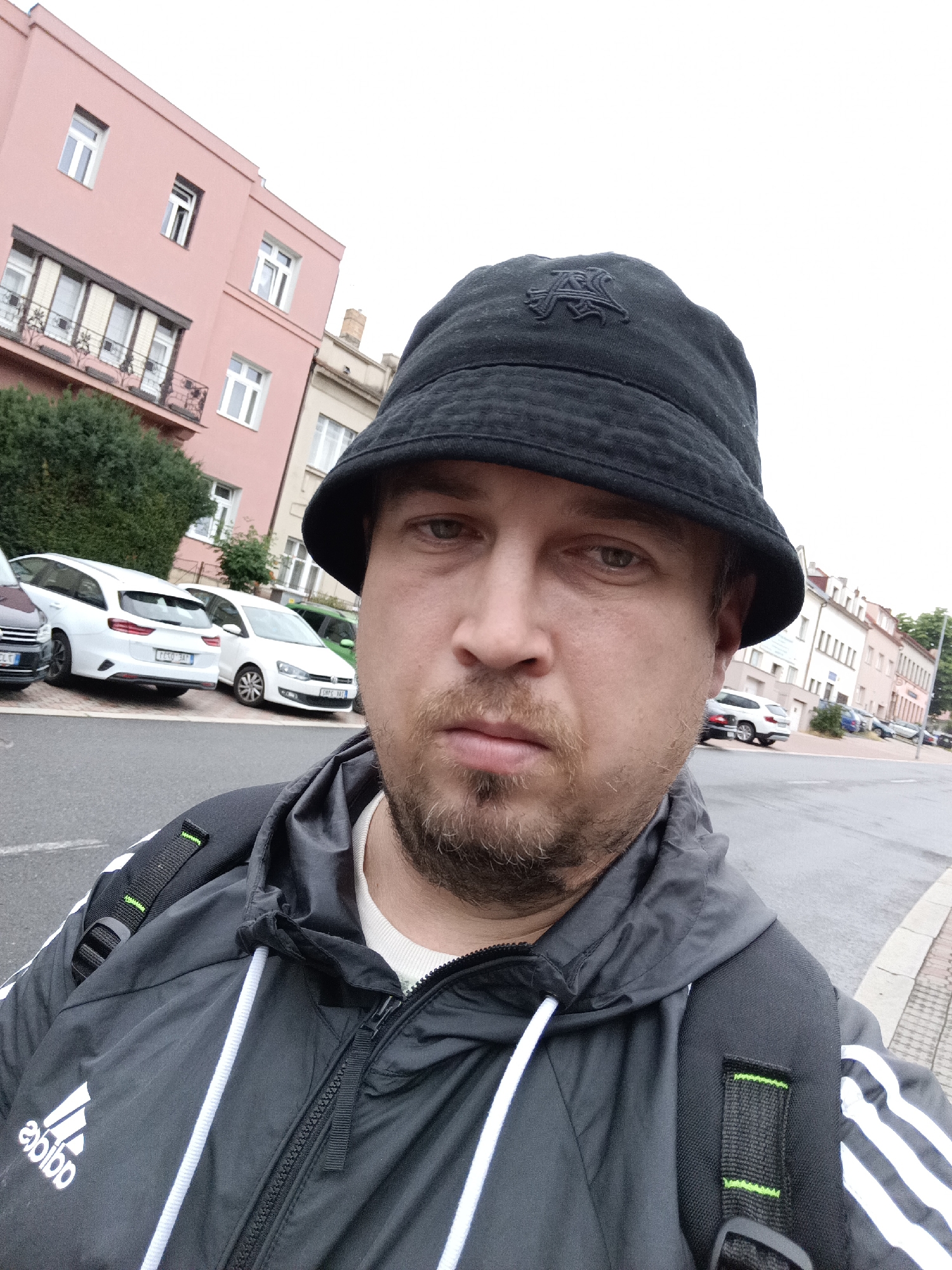 A man wearing a black bucket hat and a black Adidas jacket with white stripes on the sleeves is taking a selfie outdoors. He has a light beard and is looking directly at the camera with a neutral expression. He is wearing a white undershirt. A black backpack with neon green accents is visible over his shoulders and jacket.
The background shows a street scene in Prague, Czechia. To the left, there is a light pink building with several windows and a balcony. To the right, a row of buildings with light-colored facades and windows lines the street. Several white cars are parked along the curb in front of the buildings. The weather appears overcast, suggesting it might be daytime. The man is positioned in the foreground, with the street and buildings forming the background.