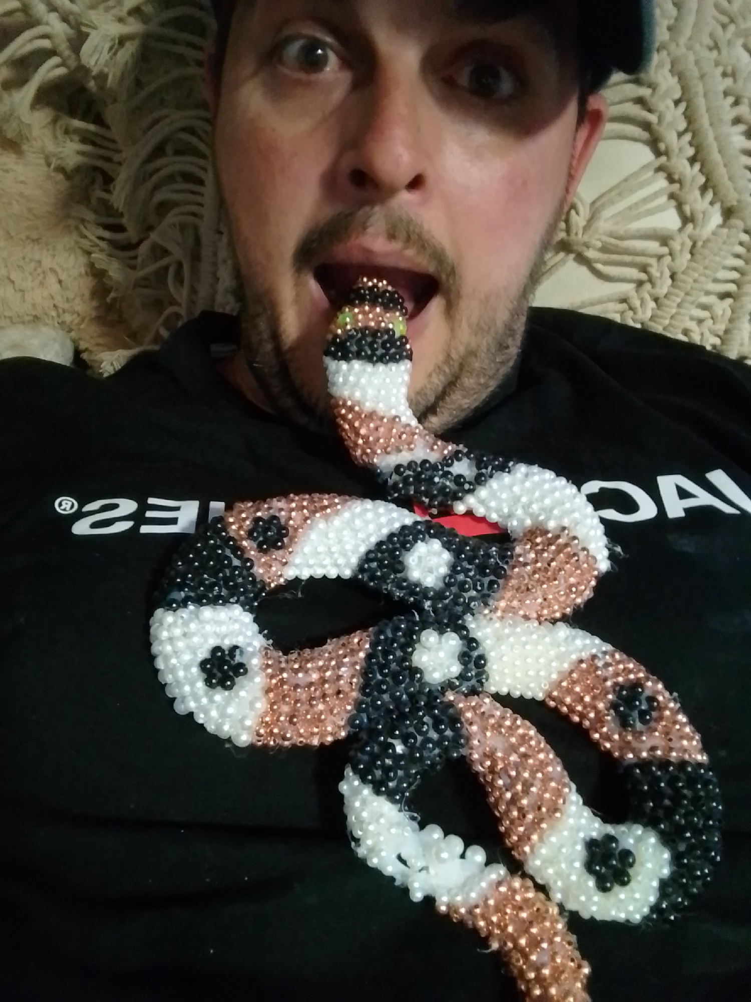 A man wearing a black t-shirt and a dark cap is lying on a textured, cream-colored couch. He is looking at the camera with his mouth open, and a beaded snake toy is partially inside his mouth. The snake is coiled around his neck and chest, with segments of black, white, and copper beads. The t-shirt has white lettering on the left side, which appears to be a logo or brand name. The background consists of the textured couch and a light-colored wall or fabric. The image quality is moderate, with some blurriness and graininess. This image is not associated with the city of Wedel, Germany.