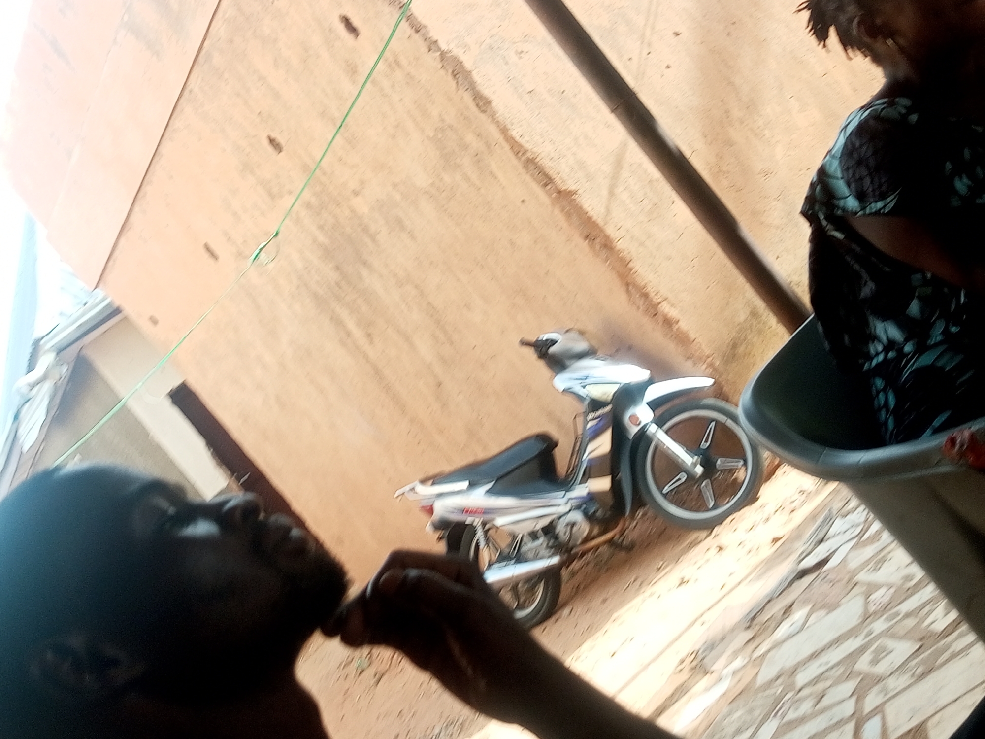 The image captures a scene in an urban environment, likely a residential area, of Unknown, Unknown. An adult individual is present in the foreground with their face partially visible and looking upwards, while another person, possibly a child, stands beside them. A washing basin or container is positioned close to the second person. In the background, a white and blue moped or motorcycle is parked near a tan-colored wall. The ground appears to be made of paving stones. A green wire is visible running diagonally across the scene. The overall perspective of the shot is tilted.