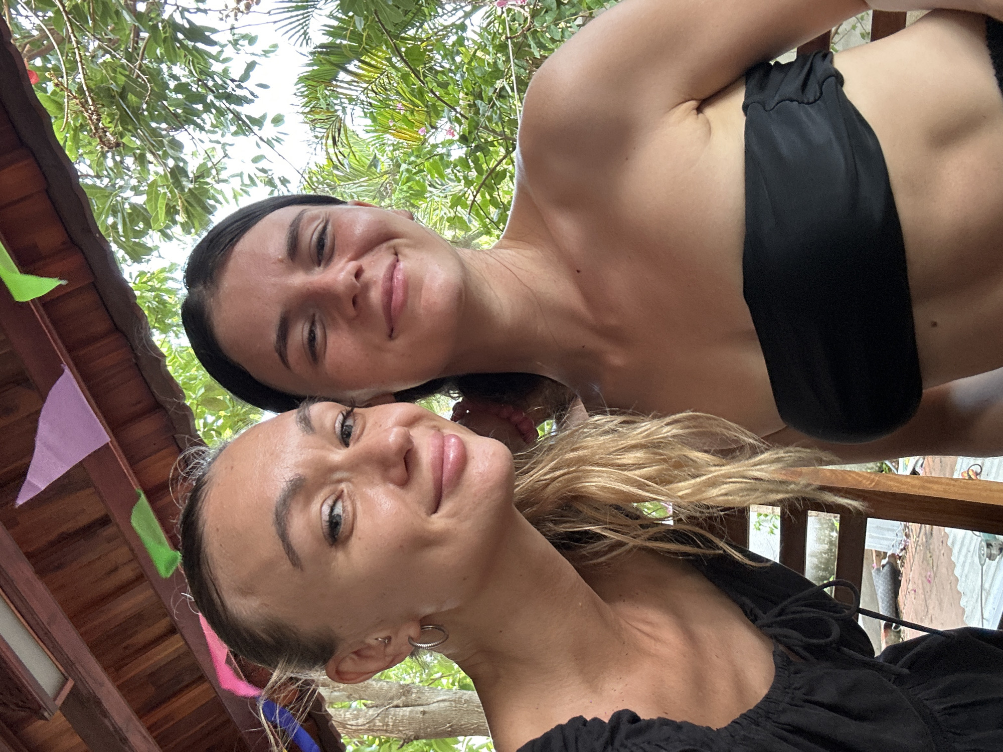 Two women pose in a selfie outdoors in Mũi Né, Vietnam. The woman in the foreground has blonde hair, and a black top with a tie, while the woman in the background has dark hair and wears a black strapless garment. They are under trees and a structure with flags. A wooden railing is also visible.