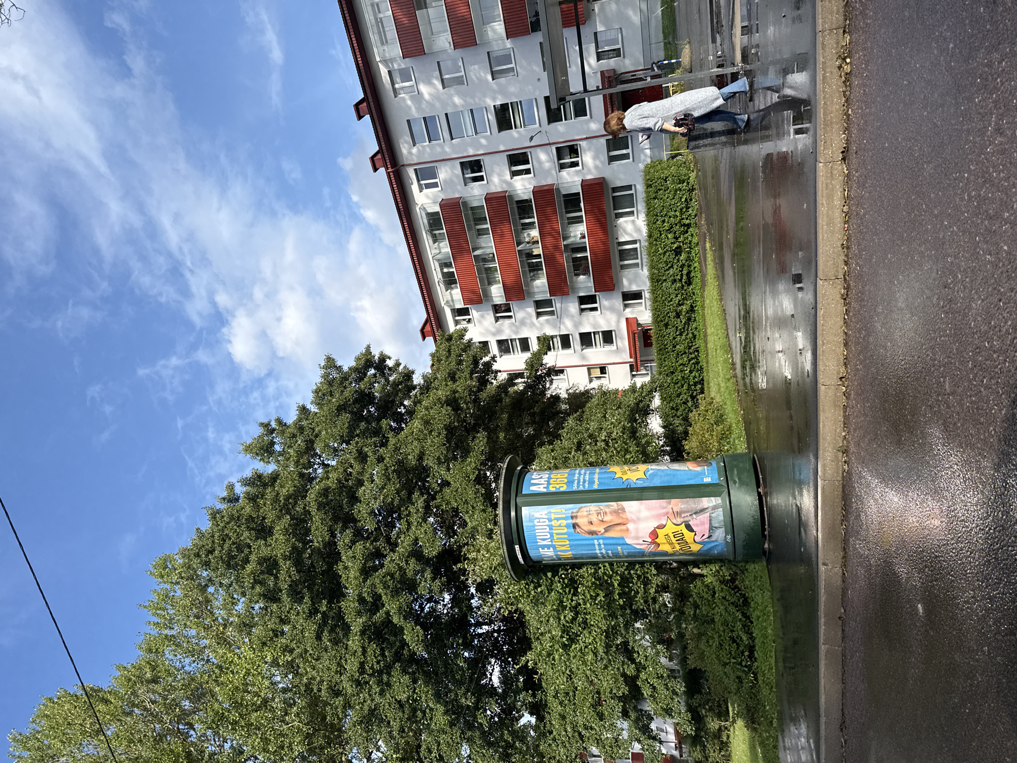 The image depicts an exterior scene, likely in a residential area, with a modern apartment building and a large tree dominating the background. The building is a multi-story structure with white facades and red balconies. In the foreground, a wet asphalt road and sidewalk suggest recent rainfall. A cylindrical advertising column stands near the tree, displaying a poster featuring a smiling woman. A person, likely a woman, walks away from the viewer on the wet sidewalk, carrying a bag. The sky is blue with scattered clouds, and the sunlight indicates it is daytime. Based on the presence of Finnish text on the advertisement column, the scene is likely located in Tallinn, Estonia, as Finnish is spoken in the Helsinki metropolitan area and is a neighboring country.