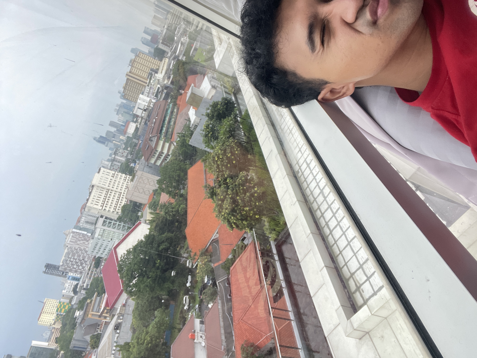 A man is seen in the foreground, looking out from what appears to be a high-rise building. He is wearing a red shirt. Beyond the window, the cityscape reveals dense urban sprawl. The buildings and structures suggest a high-density area with a mix of residential and commercial properties. The prevalence of red tile roofs indicates a tropical environment. Based on the architectural style and roof types, it is highly probable that the location is in Central Jakarta, Indonesia. The overall impression is one of a bustling urban environment.