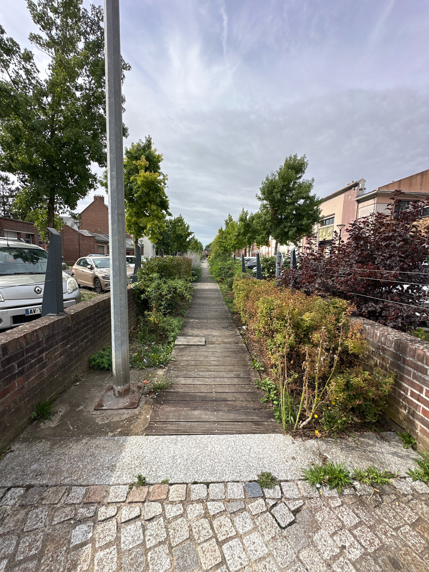 This is a ground-level, medium-shot, outdoor photograph taken on a cloudy day, likely in the city of Roye, France. The scene depicts a pathway bordered by greenery and buildings.
In the foreground, a textured cobblestone path runs diagonally from the bottom right to the bottom left. To the left of the cobblestones, a concrete sidewalk meets a low brick wall. Behind the brick wall, on the far left, is the front of a silver van with a license plate reading "AV-1". Adjacent to the van, and slightly further back, is a beige car. A tall, octagonal grey metal pole stands prominently in the left-center of the image, rooted in a concrete base.
The pathway continues into the midground as a wooden boardwalk, running parallel to the brick wall on the right. This boardwalk is lined on the left by a narrow strip of sparse vegetation and on the right by a dense arrangement of bushes and small trees with dark red and green foliage, bordered by another brick wall. Beyond this, a line of evenly spaced green trees forms a boundary along what appears to be a street.
In the background, two- and three-story buildings with light-colored facades and visible windows are visible, suggesting a residential or commercial area. The sky is overcast with scattered clouds. The overall impression is of an urban pathway with well-maintained landscaping and architecture.
