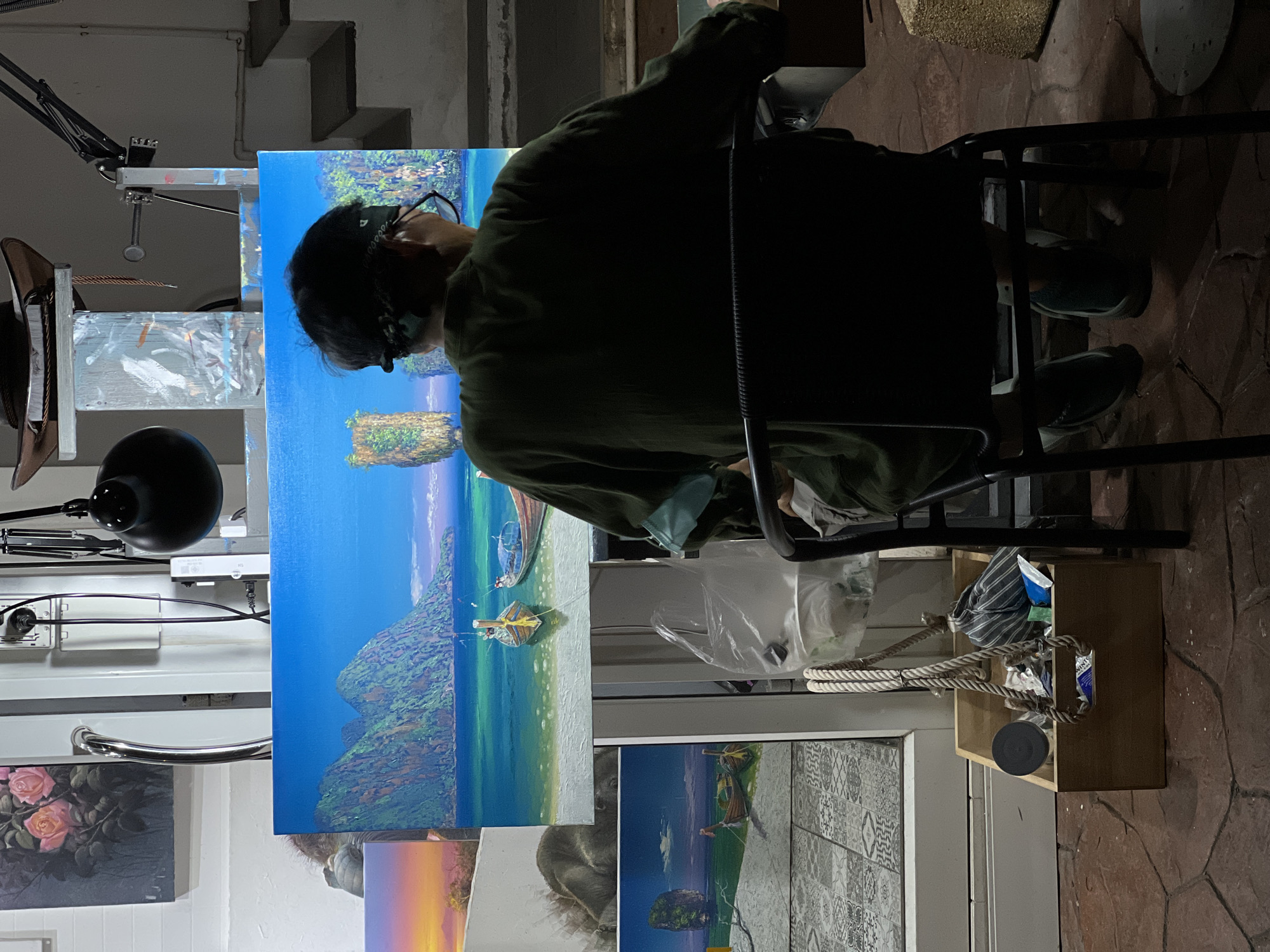 The image shows an individual seated in front of an easel, apparently working on a painting. The individual is wearing glasses and a dark-colored head covering, and is draped in a dark green garment. They are seated in a black chair. The easel supports a painting depicting a coastal scene with blue waters, a rocky outcrop, and boats on the shore. The painting is placed in a studio-like setting, with other artworks visible in the background, including a painting of flowers and a painting of a koala. There is also a toolbox or container holding various items. The scene is indoors, with a set of steps in the background. The location is Unknown, Unknown.