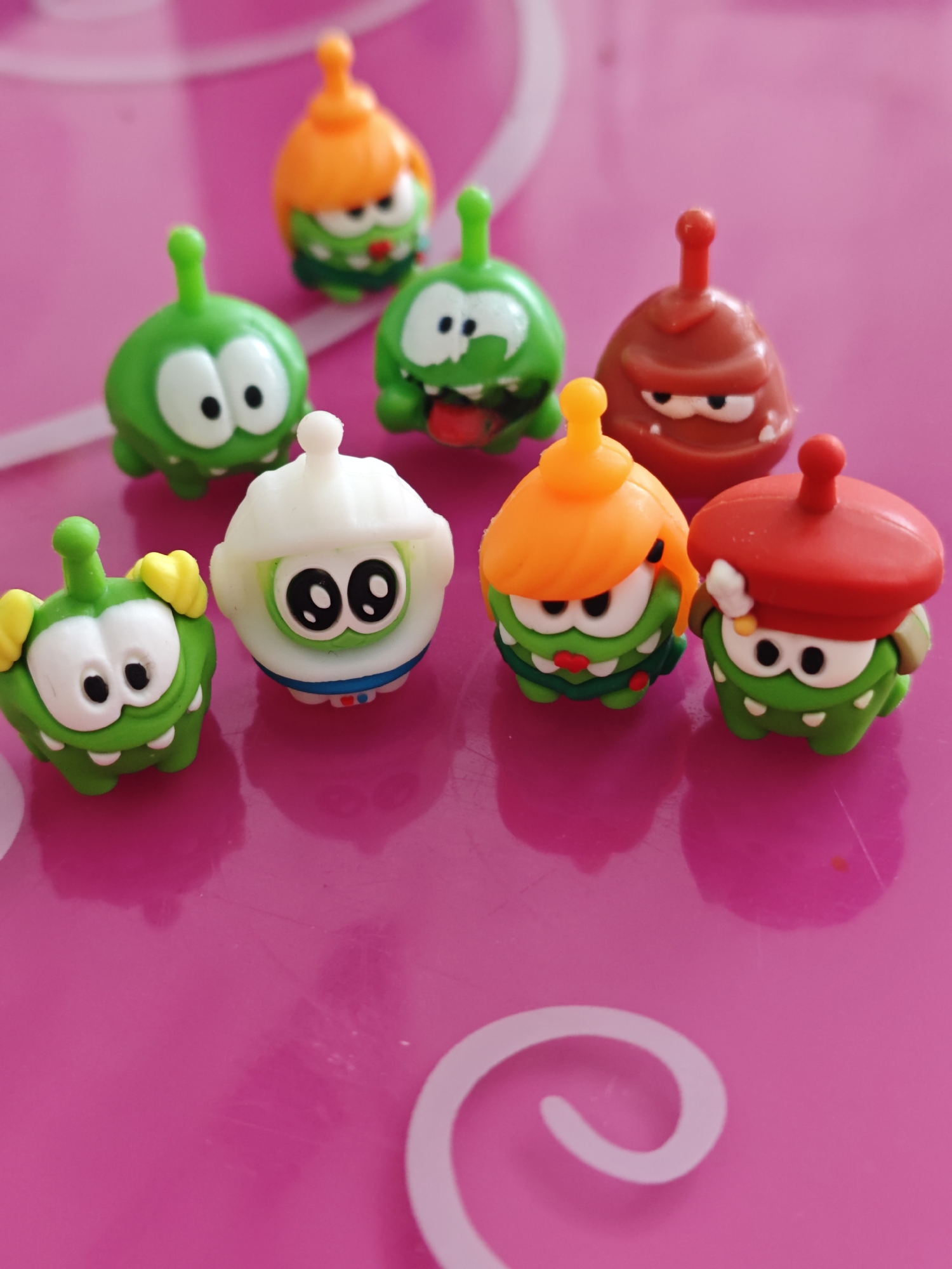 The image shows seven Om Nom toy figurines on a glossy pink surface with a white swirl design. The figurines are various colors including green, red, orange and white. They all have large eyes and small legs. The surface reflects the shapes of the toys. The exact location is not identifiable as Markovskii, Russia.