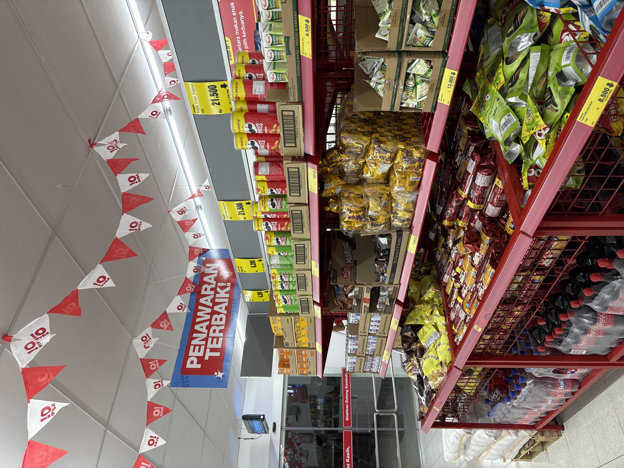 The image shows an aisle in a grocery store, likely located in Sukabakti, Indonesia, given the Indonesian language on the signage and product packaging.
The focus is on shelves stocked with various food products, including Pringles potato chips priced at Rp 21,500, Roma Malkist biscuits, and other snack items. Prices for some items such as sunflower seeds (Rp 8,500) and other products are displayed on yellow tags attached to the red shelving. There are drinks, notably Coca-Cola and Fanta, on the lower shelves.
Overhead, there are strings of pennant flags with the word "save" and the Indonesian phrase "PENAWARAN TERBAIK!", which translates to "BEST OFFER!".
A sign near the aisle entrance reads "Silahkan Datang Kembali" meaning "Please Come Again". The aisle opens to the rest of the store, seen through the glass doors at the end.