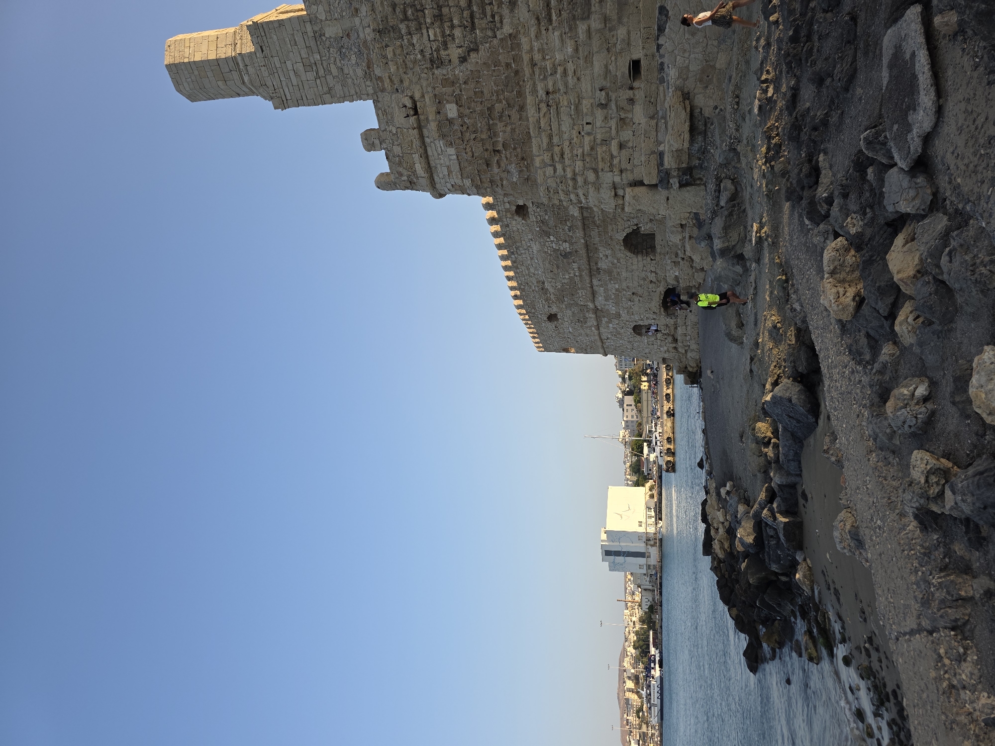The image depicts the exterior of the Koules Fortress in Heraklion, Crete, Greece. The stone structure, a prominent landmark of the city, is visible in the foreground, occupying the right side of the frame. The fortress is constructed from large, rough-hewn stones and features crenellations along its upper edge. Sunlight illuminates the stone walls, casting shadows and highlighting the texture of the masonry.
In the background, a body of water, likely the Aegean Sea, laps against a rocky shoreline in front of the fortress. Beyond the water, the cityscape of Heraklion unfolds, with buildings of varying sizes and architectural styles. A large, modern white building with blue accents stands out amongst the other structures, near the harbor. Several boats, including ferries and yachts, are docked in the harbor. The sky above is a clear, pale blue, indicating fair weather.
Two individuals are present on the rocky area in front of the fortress. One, wearing a bright yellow and green shirt and dark shorts, is walking away from the viewer. Another person, a woman in a white top and a patterned skirt, is walking towards the right side of the frame, away from the fortress. The scene is set during what appears to be late afternoon or early evening, with the sun casting long shadows. The overall impression is one of historical grandeur meeting modern port life under a clear sky.
