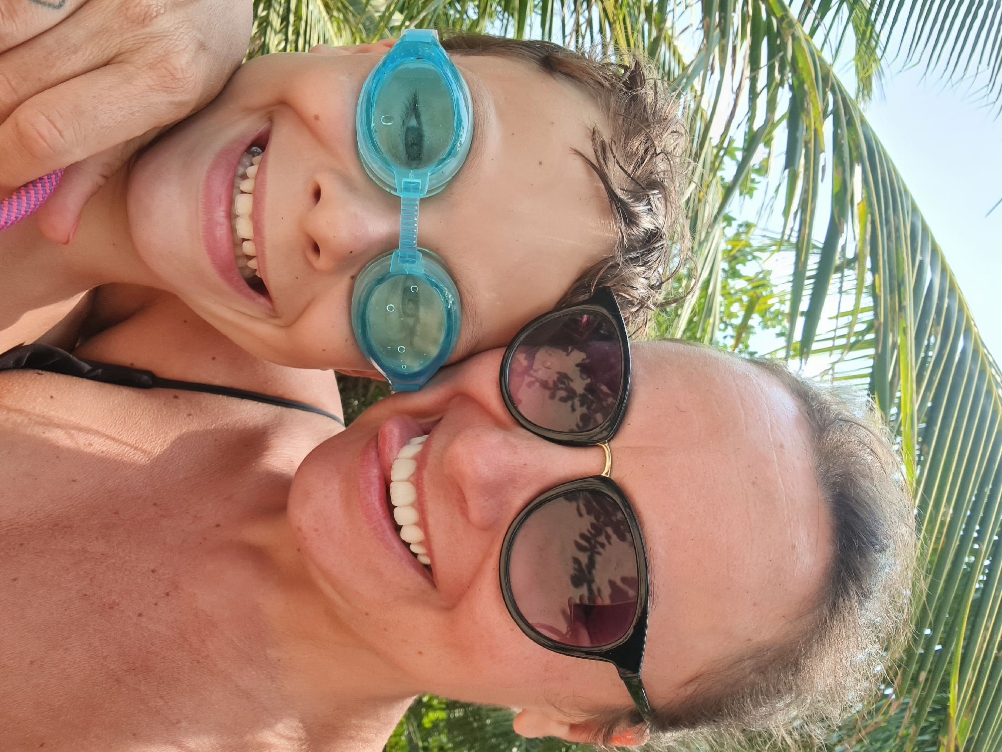 Two individuals, a child wearing blue goggles and an adult wearing sunglasses, are cheek-to-cheek, smiling and facing the camera. They appear to be in a tropical environment, indicated by the presence of palm fronds visible in the background and reflected in the sunglasses. It is possible that the setting is in the city of Đà Nẵng, Vietnam, based on the tropical climate and common presence of palm trees.