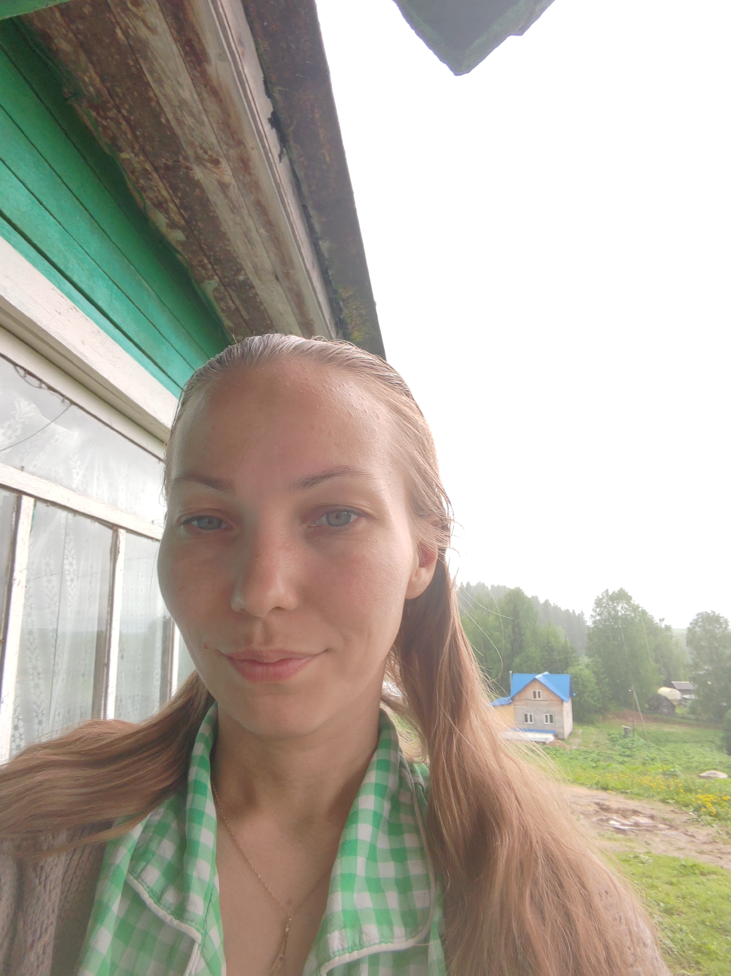 A fair-skinned woman with blonde hair pulled back into pigtails is the primary subject of a selfie taken outdoors in what appears to be a rural area. She is wearing a green and white checkered shirt over a neutral-colored sweater, and a necklace with a charm is visible.
The background shows a building with green siding and white-framed windows to the left of the woman. In the distance, another building with a blue roof is visible, along with greenery and trees.
The selfie is taken on what seems to be a foggy or overcast day in Чухлэм, Russia.