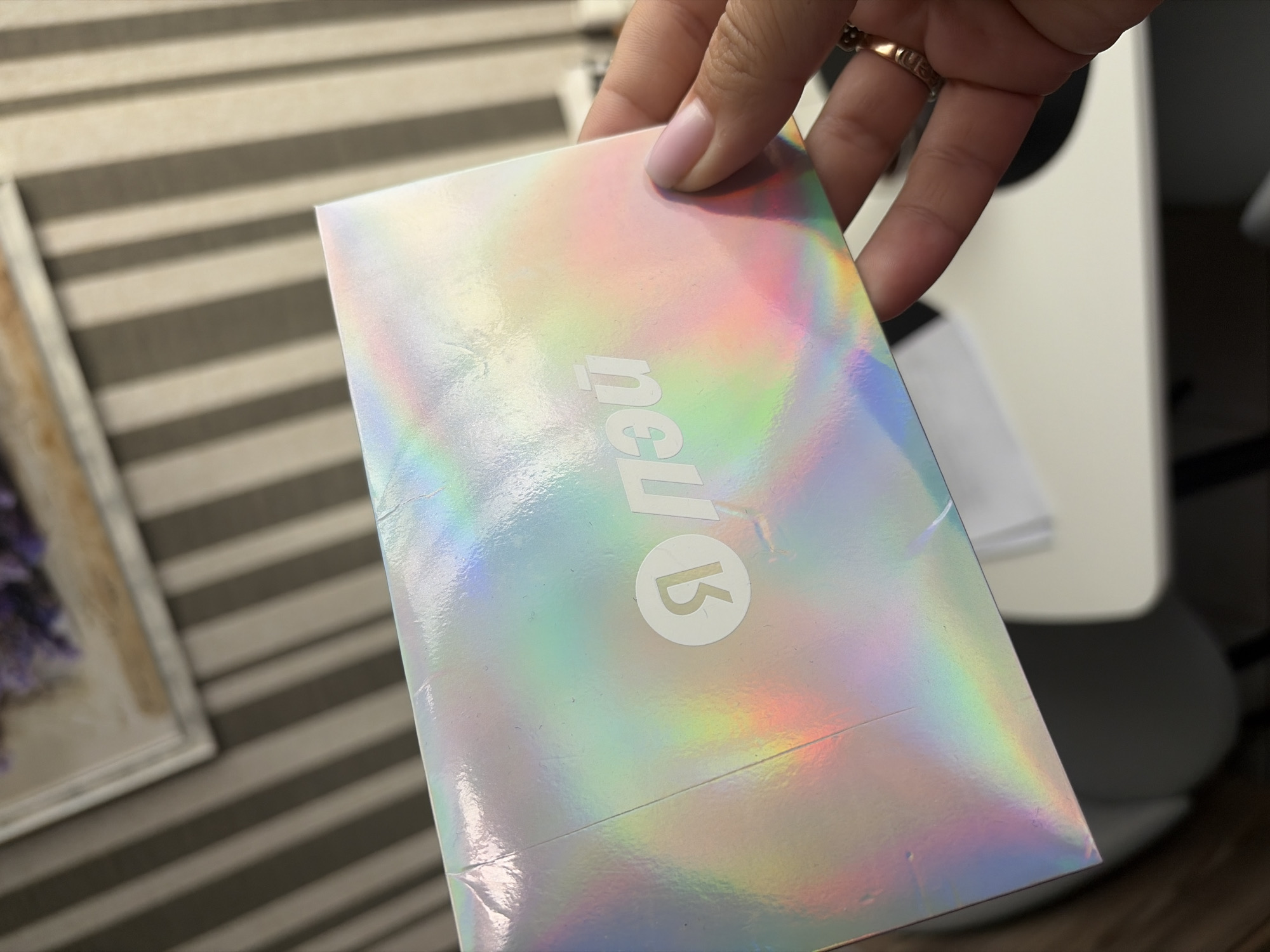 A person's hand is holding a holographic envelope. The envelope has a circular logo with a stylized letter "Я" and the Cyrillic letters "ПЭЙ" printed on it. The background appears to be a wall with vertical striped wallpaper and a framed picture hanging on it. The scene is indoors. There is no information about the location in Sochi, Russia.