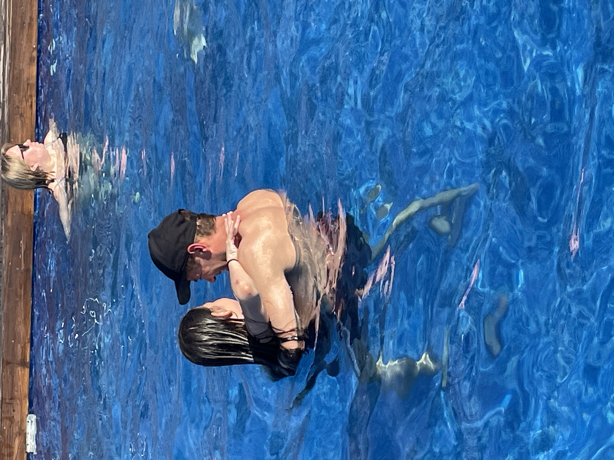 The image depicts a couple embracing in a swimming pool. A man with a baseball cap is holding a woman with long dark hair closely, both submerged in the water. The water's surface is rippled. Another woman is seen in the pool near the edge. The location, Люкшудья, Russia, cannot be confirmed from the image.