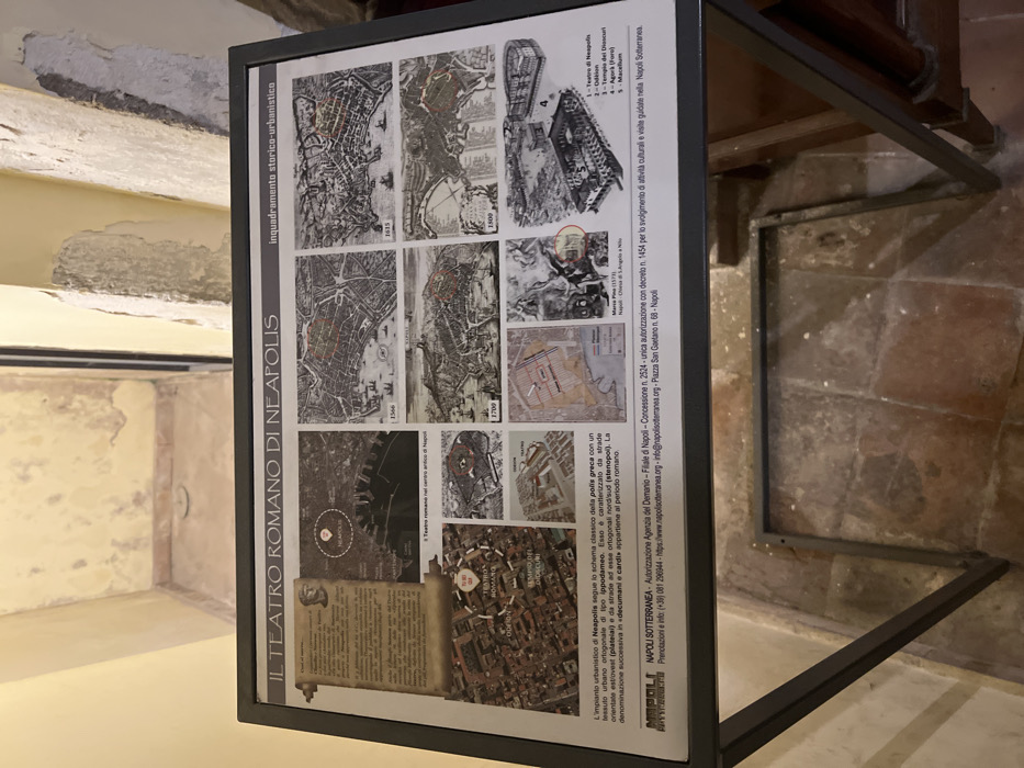 The image shows a black rectangular informational sign with a white background and the text "IL TEATRO ROMANO DI NEAPOLIS" printed on it. The sign is on a stand with a red tile floor. The sign is positioned near a wall. The sign has an array of black and white maps showcasing the evolution of the city of Naples, Italy, over several centuries. The location of the sign is in the city of Naples, Italy at the coordinates 40.852 latitude and 14.257 longitude. The sign is mounted on a stand with a red tile floor. There is a black framed doorway in the background.