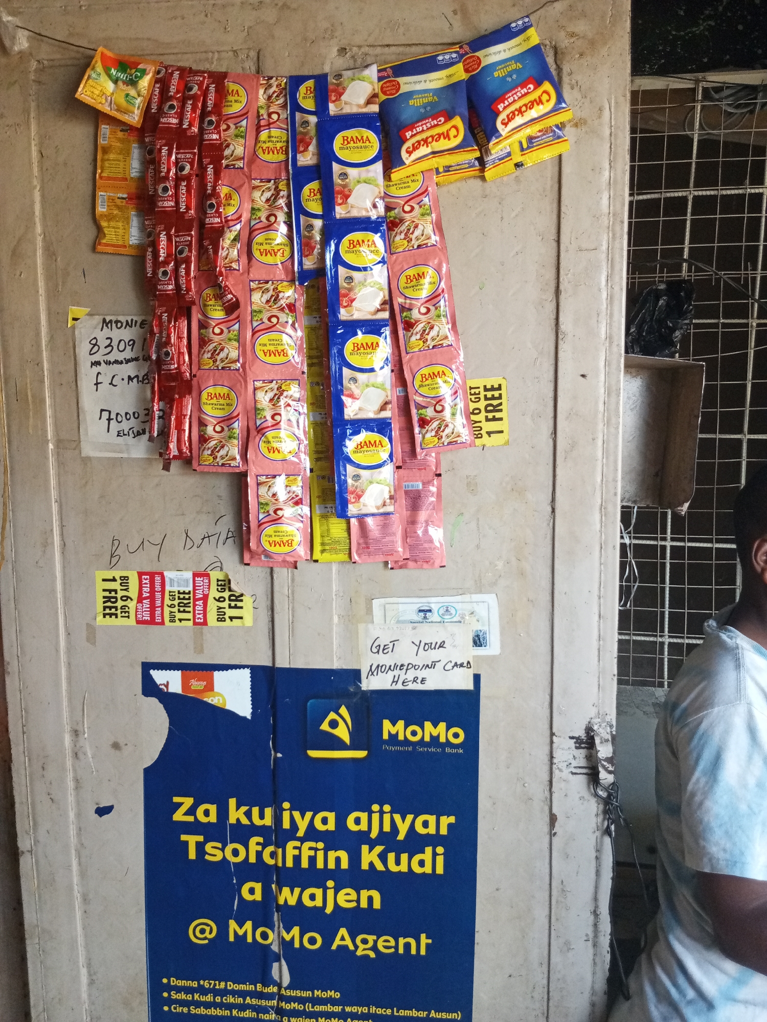 The image shows a close-up of a weathered wooden door with various items attached to it. On the upper part of the door, there are numerous sachets of food products, including Nescafe, Bama Mayonnaise, and Checkers Custard. Below these, a handwritten note is visible, along with some yellow promotional stickers. Further down, a large blue poster with yellow text and logos related to "MoMo Payment Service Bank" is affixed to the door. The door itself is aged and shows signs of wear and tear, with peeling paint and some metal fixtures. A portion of a person's shoulder and arm, wearing a tie-dye shirt, is visible on the right side of the frame, suggesting they are standing next to the door. The environment appears to be an outdoor setting, possibly a shop or market stall, given the display of products.