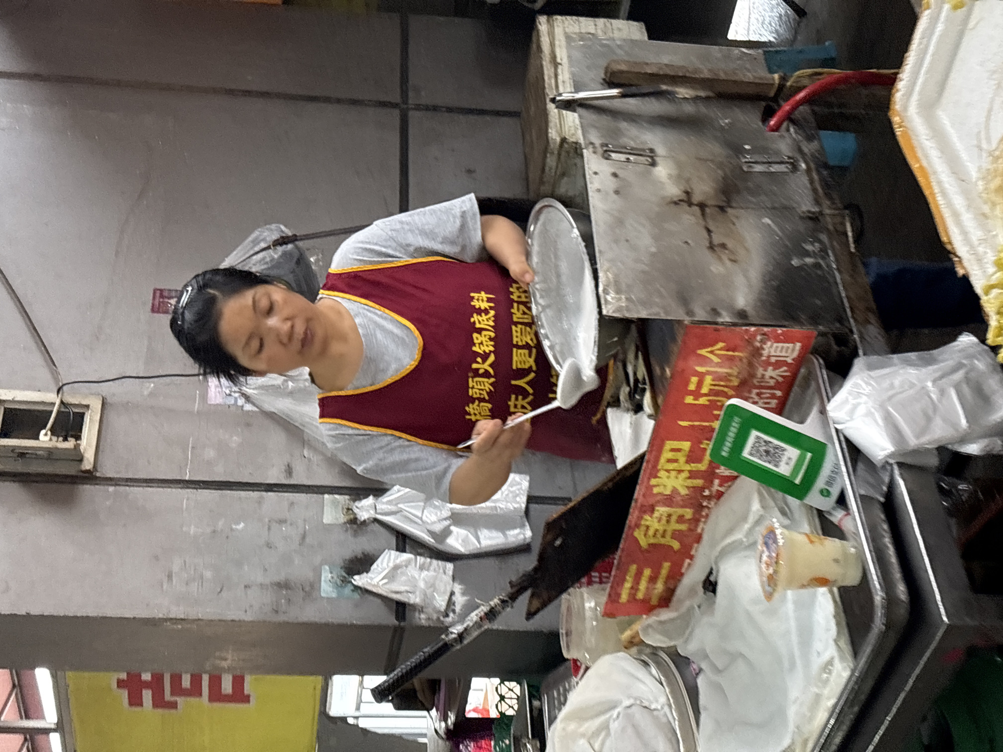 Here is a description of the image:
A woman with black hair and a red apron is standing at a food stall. She is holding a round, silver plate in her left hand and using a long, thin utensil in her right hand to move something white from the plate. The stall has a stainless steel counter with a small wooden block on top. There are various objects scattered around the counter, including a red sign with Chinese characters, a green QR code, and some plastic bags. The background consists of a concrete wall. The stall is located in Jiangjin, China, where the sign on the apron, "Qiaotou Hot Pot Ingredients," is a popular local dish.