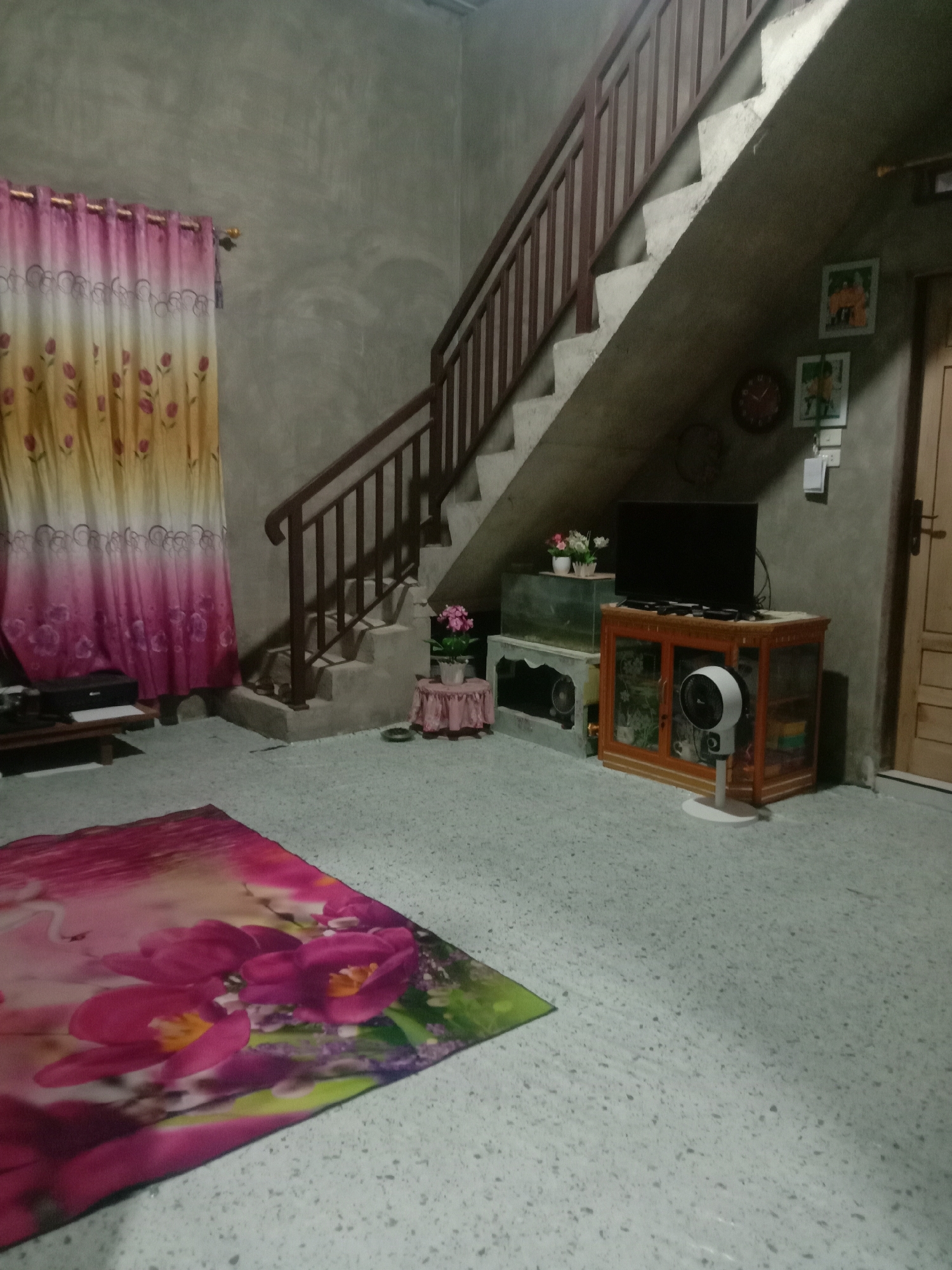 The image depicts the interior of a room, possibly in Lhokjok, Indonesia, judging by the vernacular architectural style. The room is dimly lit and appears to be a living space. In the foreground, there is a rectangular rug with a floral pattern. Behind the rug is a low, dark-colored table, partially obscured by curtains. The curtains are patterned with pink, yellow, and white hues.
A staircase with a wooden handrail ascends along the right side of the room, partially blocking access to a door. A television is visible atop a wooden cabinet with glass doors. To the left of the cabinet is a fish tank, and a small white fan stands in front of the cabinet. A small table with a potted plant sits to the left of the fish tank. The walls are unadorned concrete, with a few small framed pictures and a clock mounted near the door. The floor appears to be tiled with a speckled pattern. There is no indication of other people or activity in the frame.