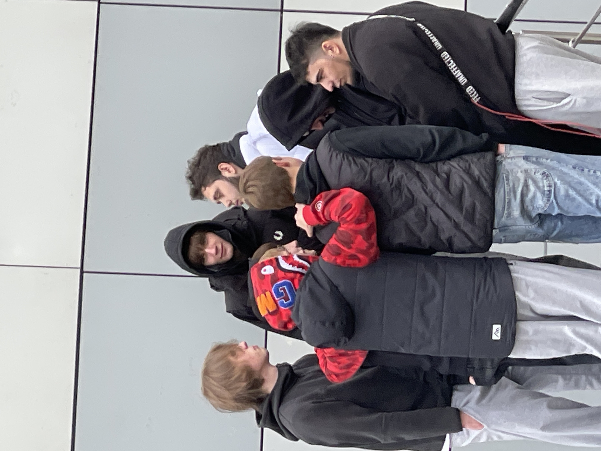 Seven young men are standing closely together indoors against a light gray wall. They are dressed in a variety of casual clothing including hoodies, jackets, and jeans. The photograph appears to be taken in a public space, potentially a transportation hub such as an airport or train station in Moscow, Russia. The men seem to be engaged in a private conversation or observation.