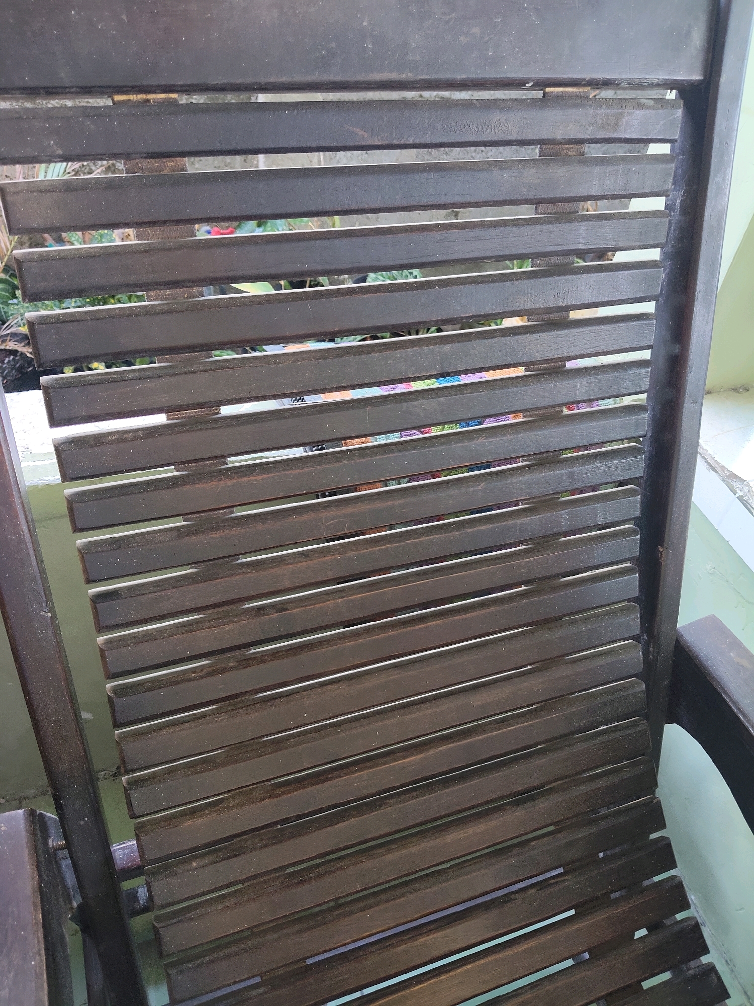A close-up, eye-level shot shows the dark brown wooden slats of a chair's backrest, with glimpses of colorful fabric and green foliage visible through the gaps. The chair appears to be made of dark wood, likely teak, and is designed for outdoor use. The slats are evenly spaced, and the wood shows signs of weathering. The background is blurred, but suggests a setting with greenery and possibly a light-colored wall or structure. Based on the visual cues, the scene is likely from a residential outdoor area in Kupang, Indonesia, possibly a veranda or garden. There are no people or specific actions depicted, focusing solely on the texture and construction of the chair.