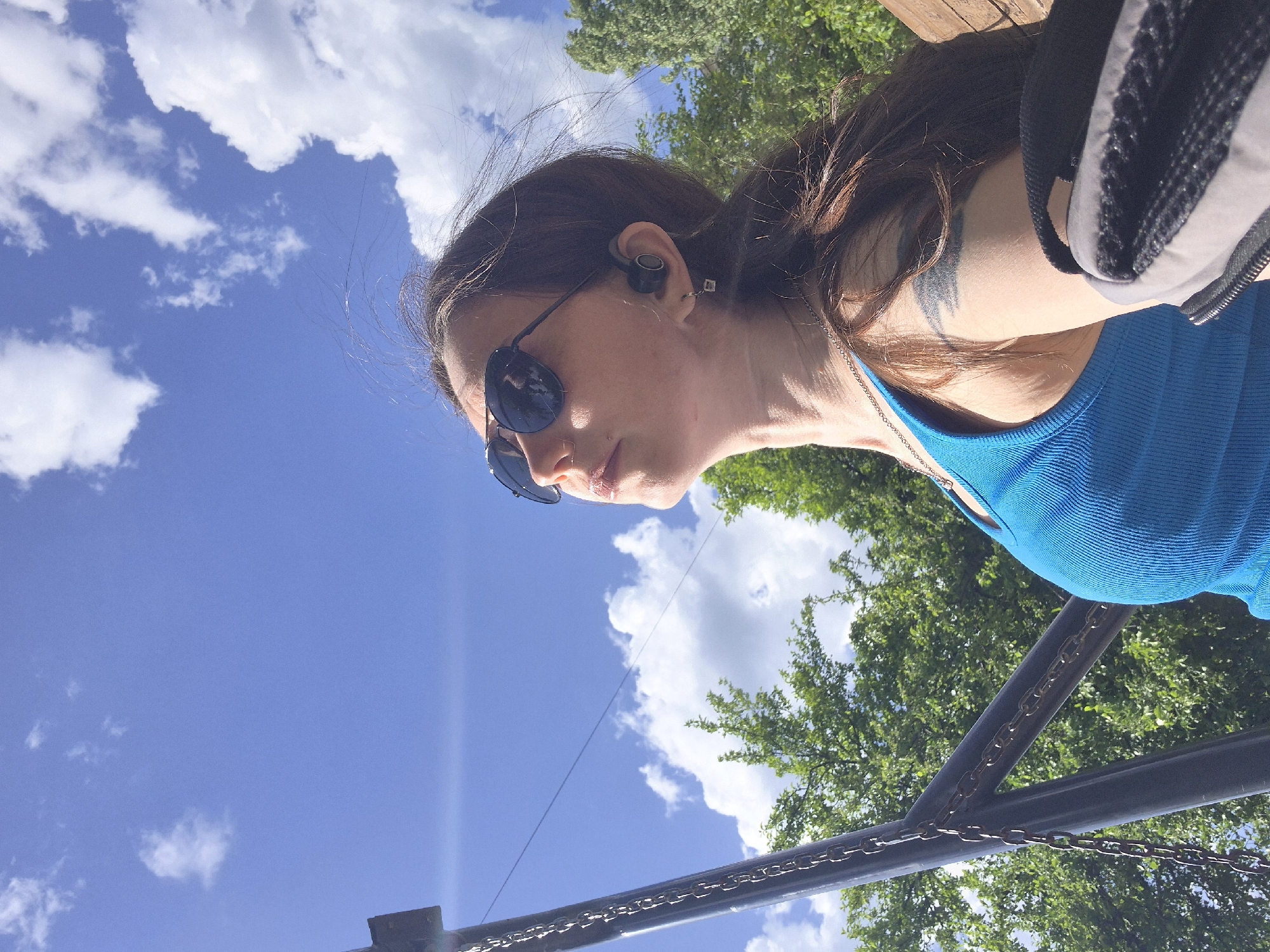 A woman with long brown hair, sunglasses, and a blue tank top is seen from a low angle under a bright blue sky with scattered clouds. She wears wireless earbuds in her ear and a silver necklace. A tattoo is visible on her neck. The background includes trees and a metal structure with chains. The location is unspecified.