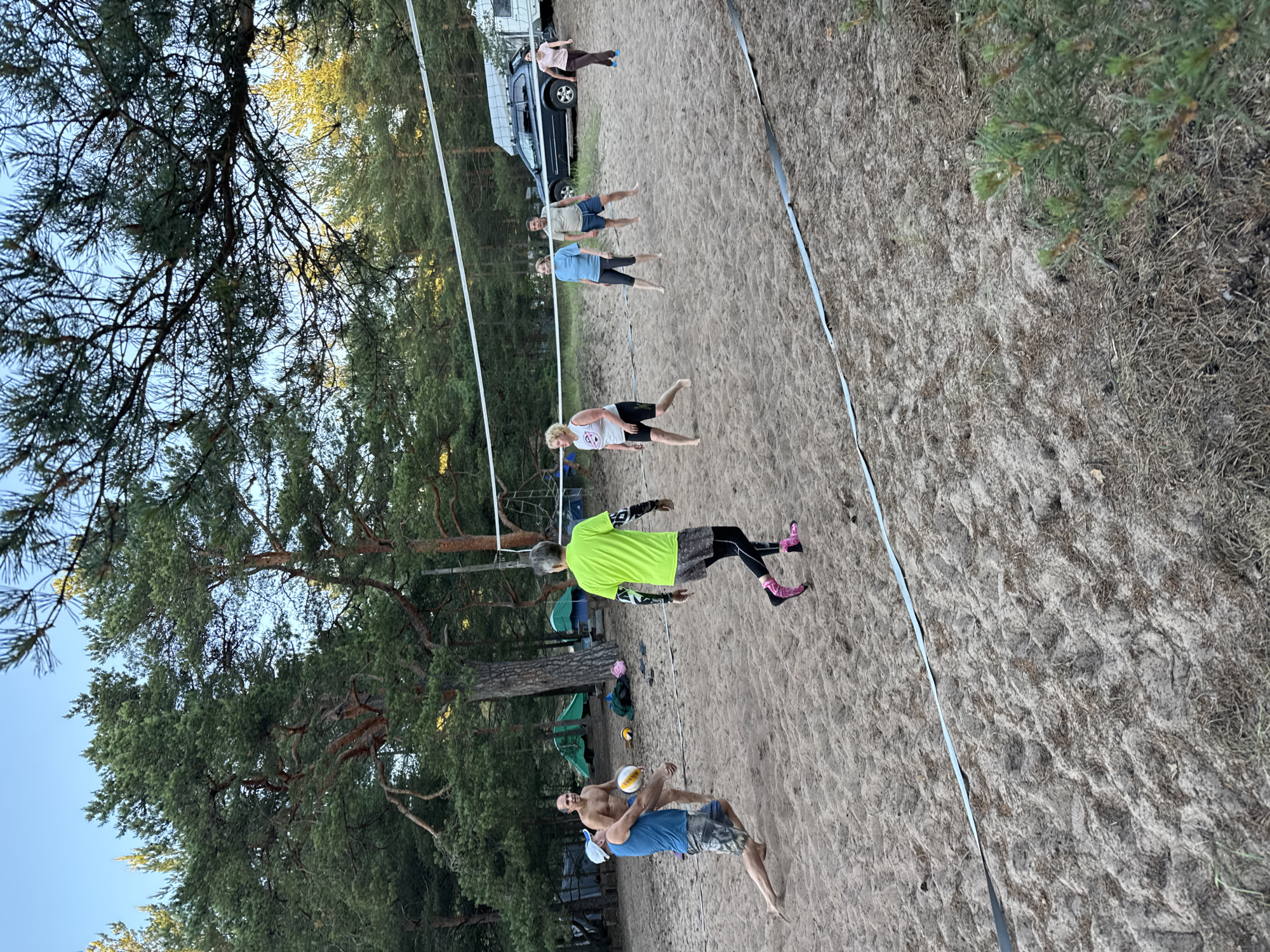 In Zelenaya Roshcha, Russia, a group of people is playing volleyball on a sandy court. The court is surrounded by pine trees, and there are tents and a vehicle in the background. A man in a blue shirt and camouflage shorts is in the foreground, jumping to hit the ball. Another man, shirtless, is standing behind him. A person in a bright green shirt is also on the court, facing away from the camera. On the other side of the net, three women are positioned on the sand. The sun is low in the sky, casting long shadows across the court.