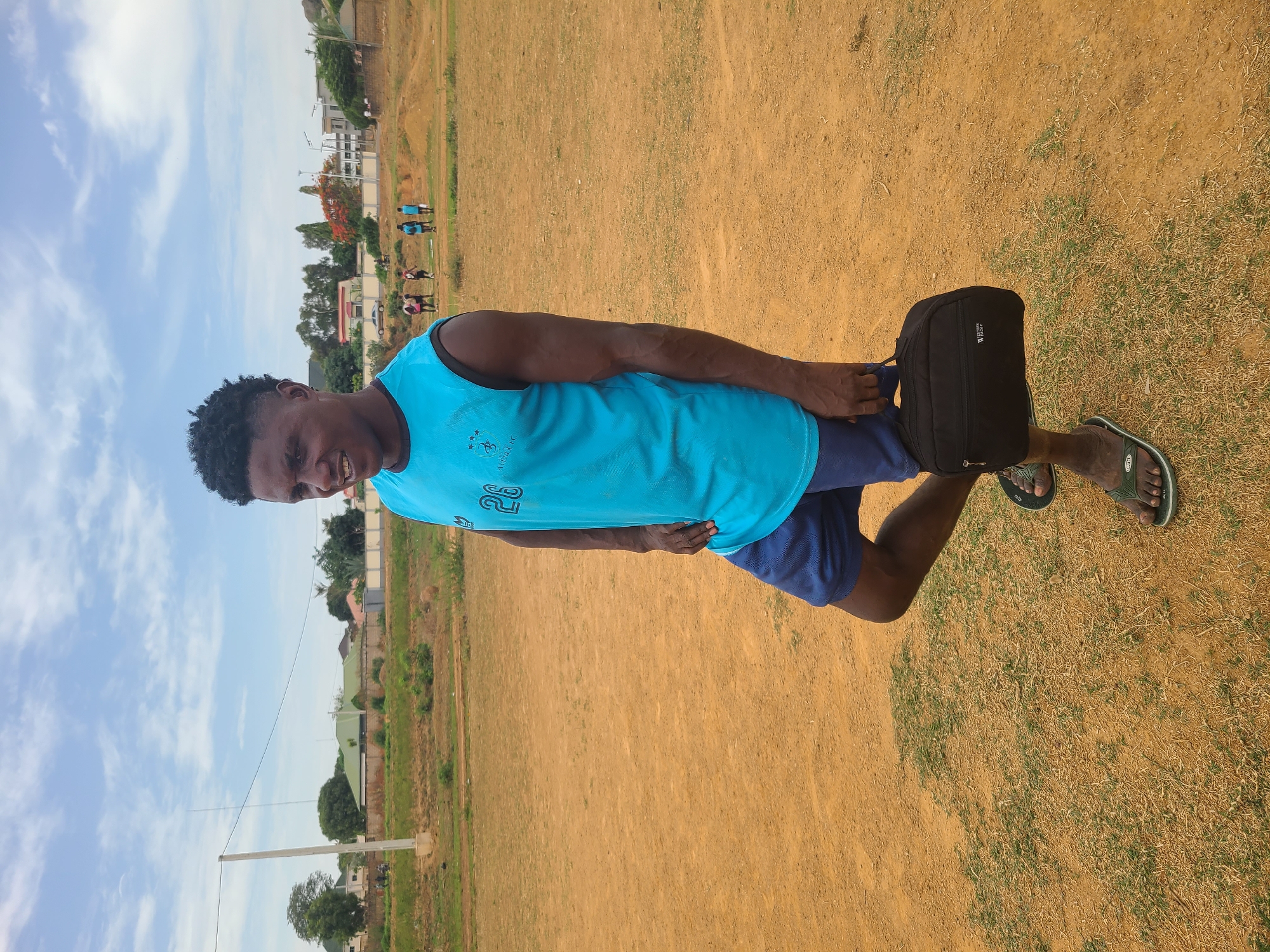 A man wearing a blue tank top and dark blue shorts stands on a dirt and grass field. He is holding a black bag near his right leg. He is wearing green sandals. In the background, there are buildings and trees. Other people are discernible in the distance. The photo was taken in Barikin Jirga Summa, Nigeria.