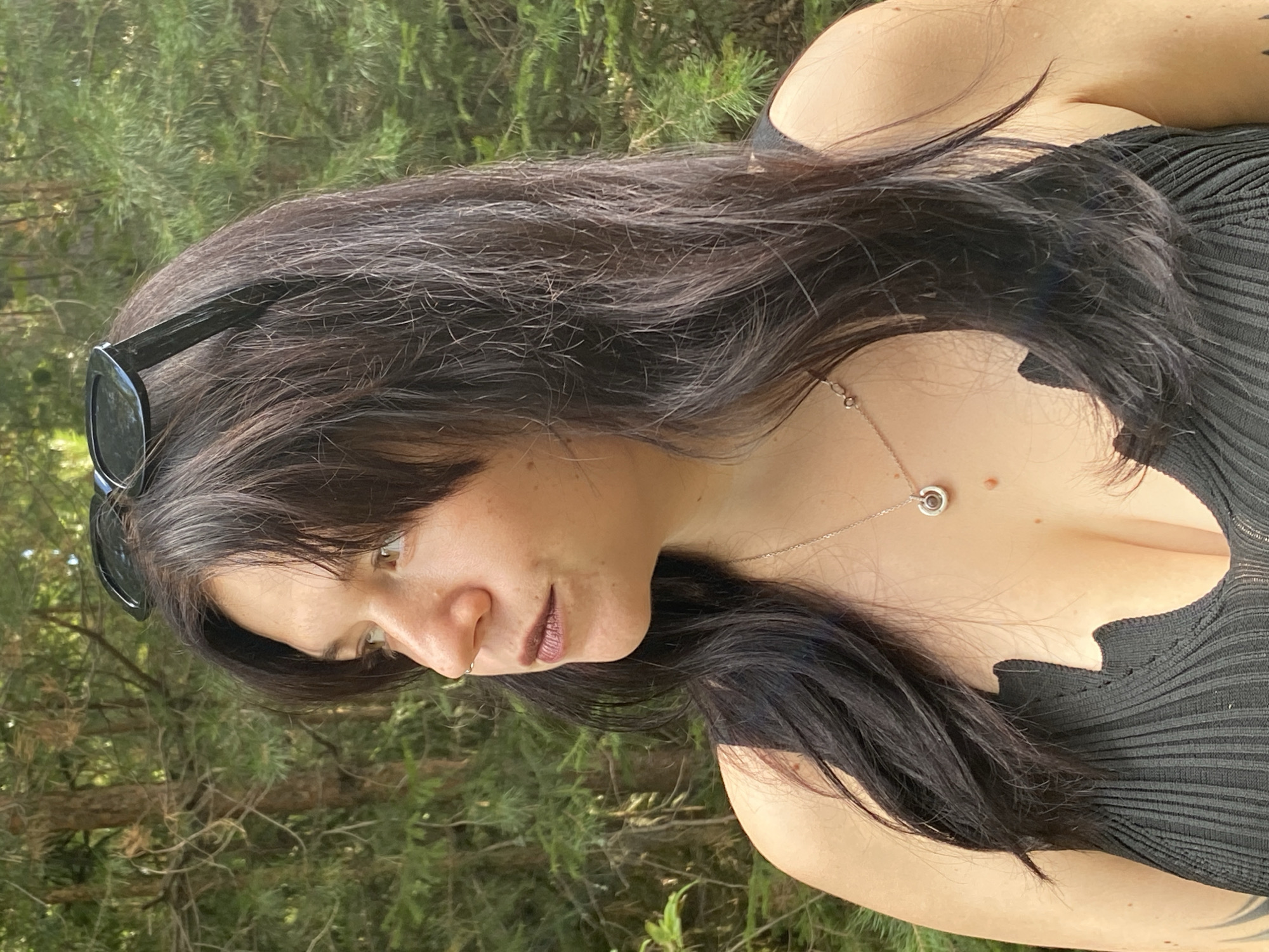 In a medium shot, a woman with dark hair and fair skin is photographed outdoors against a backdrop of trees. She is looking to her left, her head tilted slightly upward. Sunglasses are perched on top of her head, and she is wearing a black top with a scalloped neckline. A delicate necklace with a circular pendant rests on her collarbone. The setting appears to be a natural environment, possibly a park or forest, with lush green foliage and tree trunks visible behind her. There is no indication of the specific location within the city of Фоки, Russia.