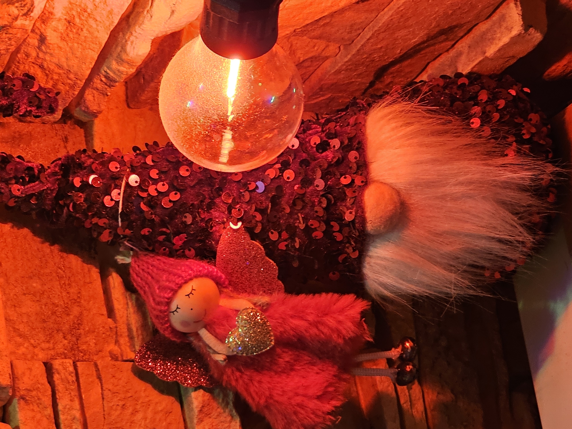 The image shows two festive decorations, possibly for Christmas or New Year, placed against a stone wall illuminated by warm orange light. On the left is a small, red-clad fairy-like doll with closed eyes and a knitted red hat. It is holding a golden heart. To its right stands a gnome-like figure, also adorned with red sequins, featuring a long white beard. In the foreground, a large, glowing lightbulb is visible, casting a bright, warm light and reflecting on the objects. The scene evokes a festive and cozy atmosphere, potentially a shop window display or home decoration. There is no visible indication of the city of Skadovsk, Ukraine.