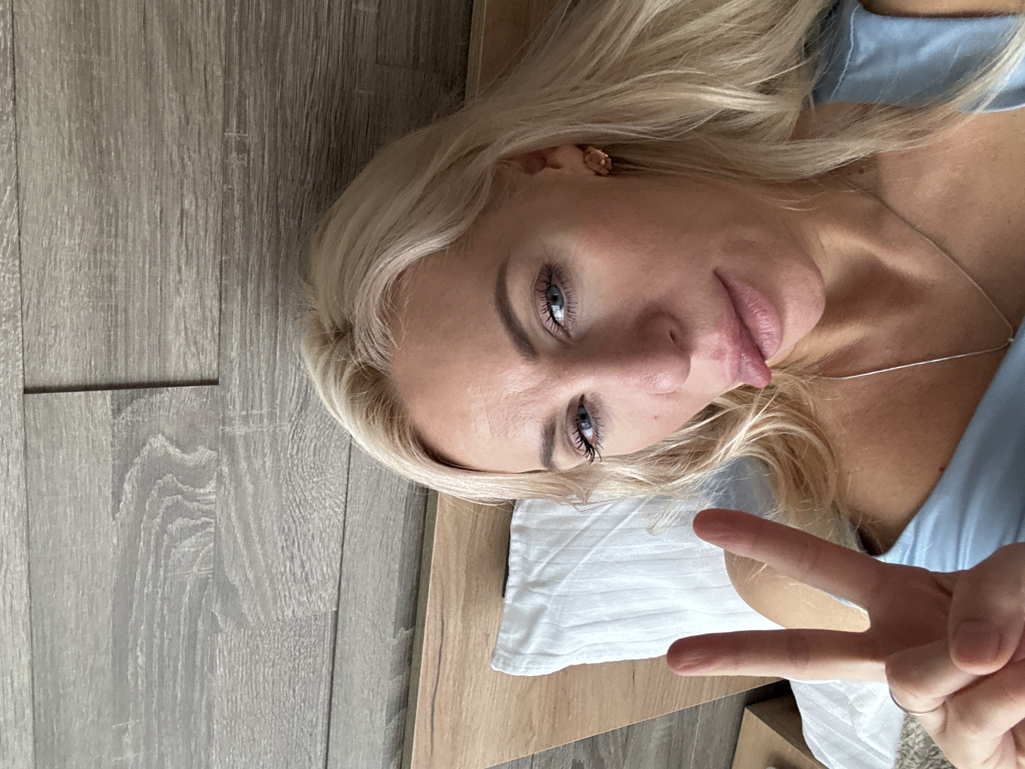 A close-up, high-angle, selfie-style photograph shows a blonde woman lying in bed. She is looking directly at the camera with blue eyes and has her tongue sticking out slightly. Her right hand is raised, displaying a peace sign with two fingers extended. The background consists of a wooden headboard and wall, and a white pillow is visible on the left side of the frame. The woman is wearing a light blue top and a silver necklace. The location is identified as Starye Chernitsy, Russia.