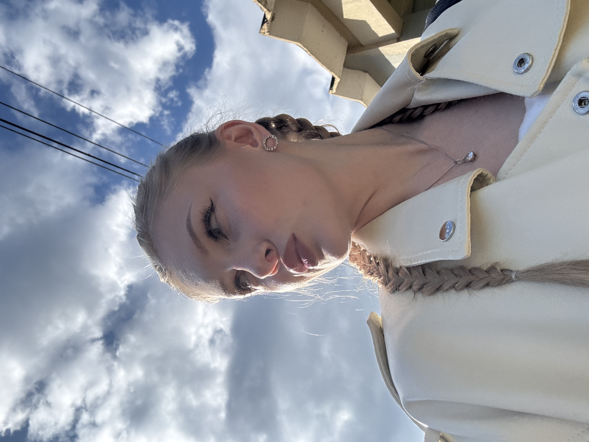 An outdoor, medium shot captures a young blonde woman from the chest up, looking to her left, with her mouth pursed. She is wearing a light-colored jacket with silver snaps and a delicate silver necklace with a small pendant. Her hair is styled in two braids, one hanging over her shoulder and the other trailing down her back. She has on a circular, sparkly earring in her visible ear. The background consists of a blue sky with white, wispy clouds and a portion of a tan building with angled roofing. Electrical wires are visible strung across the sky in the upper right quadrant of the image. The lighting suggests it is daytime. The scene appears to be in a residential or commercial area, possibly Yekaterinburg, Russia.