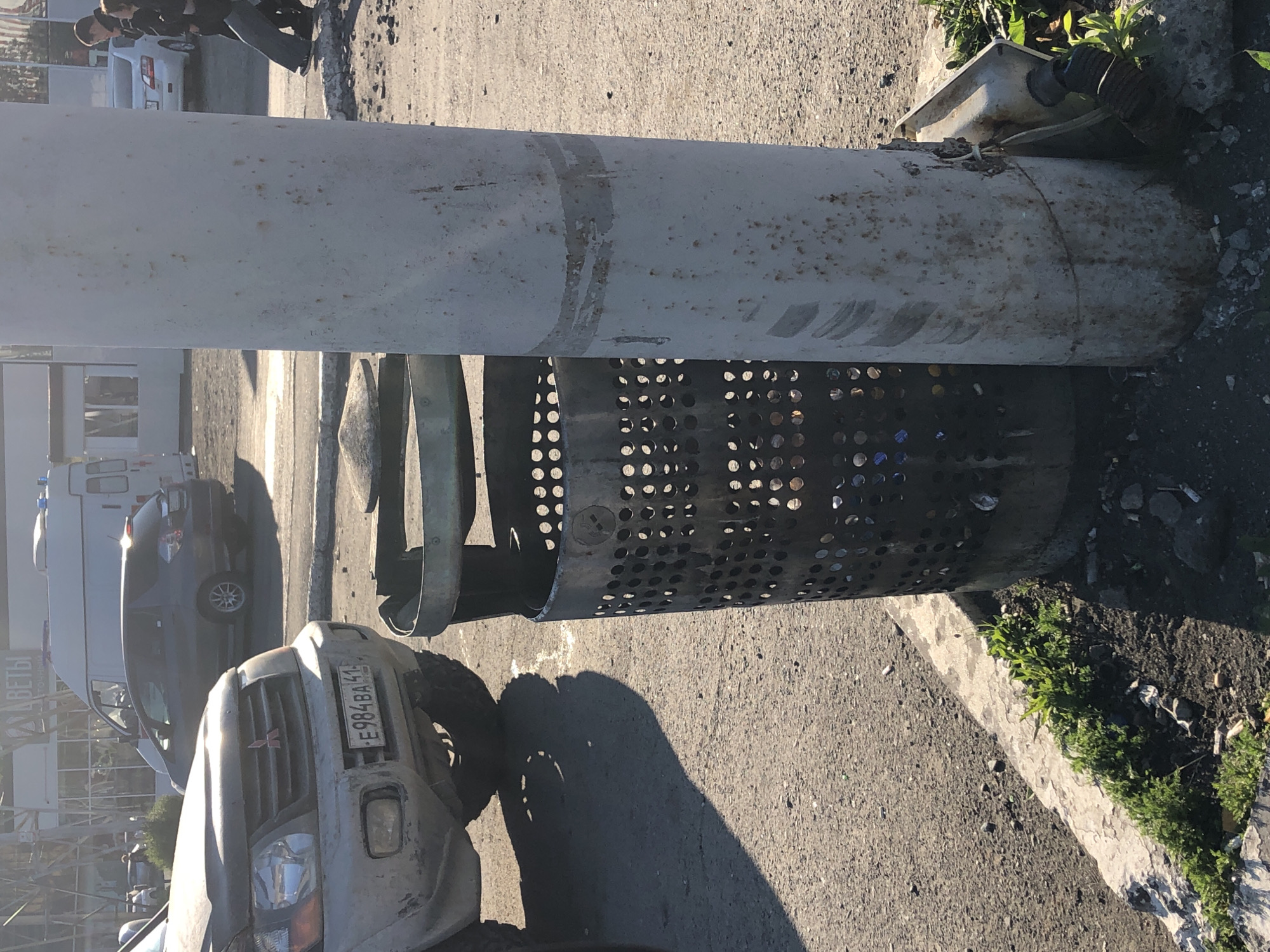 The image shows a street scene in Petropavlovsk-Kamchatsky, Russia, bathed in bright sunlight, casting long shadows. In the foreground, a metal trash can with a perforated body stands next to a light pole. The pole is weathered and discolored, with rust spots and visible scratch marks. A damaged electrical conduit is attached to the base of the pole. To the left, the front of a silver SUV is visible, its license plate reading "E984BA 41". The SUV appears dusty. Behind the SUV, a dark-colored hatchback is parked. In the background, a white van with flashing blue lights is parked, and further back, a white car is visible. Two individuals, a man and a woman, are walking away from the camera on a sidewalk in the middle ground. The ground is paved with asphalt and gravel, with a concrete curb and some sparse green vegetation growing along it.