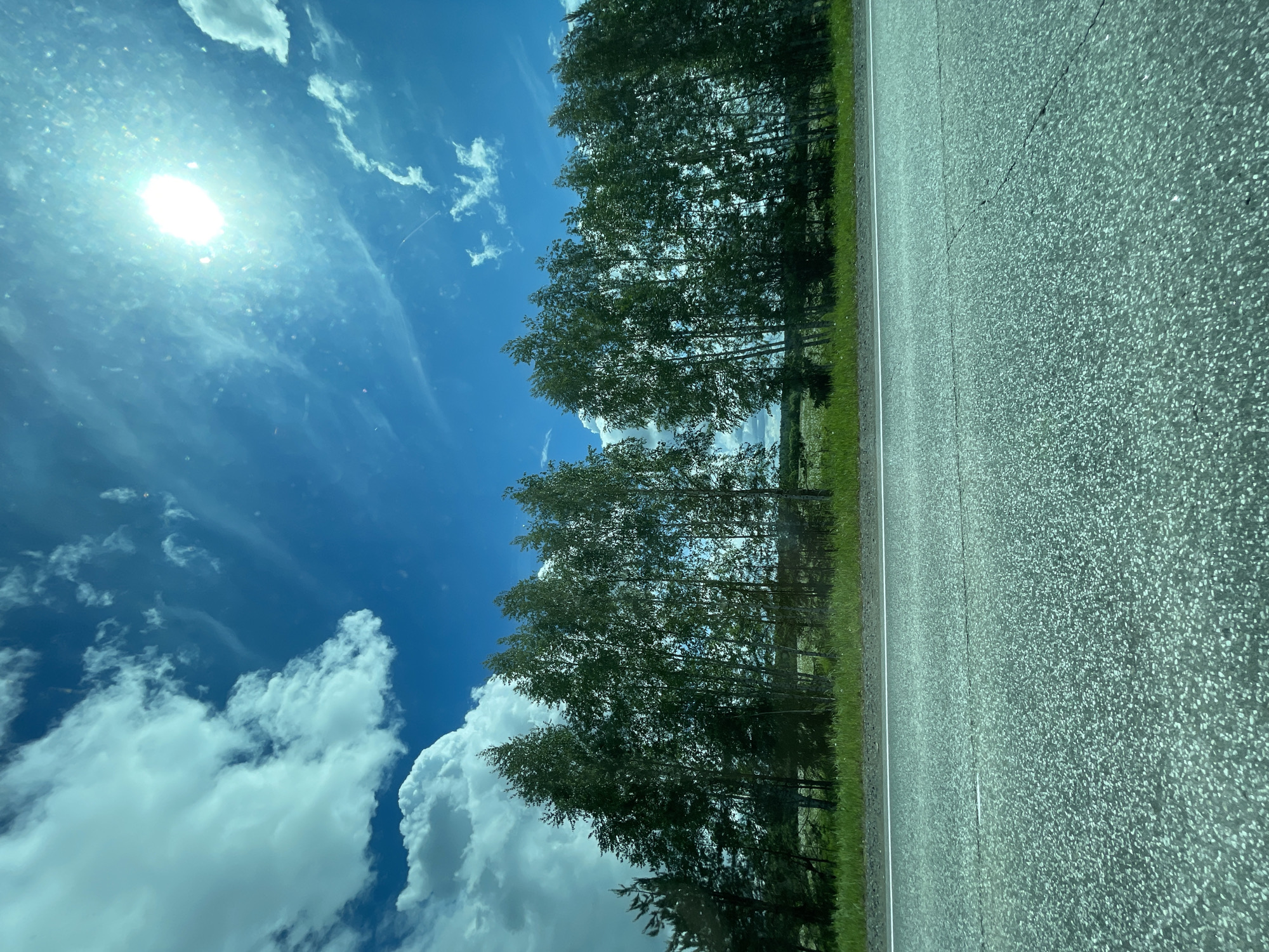 The image shows a road adjacent to a line of trees under a blue sky with clouds and a visible sun. The scene is possibly taken from inside a moving vehicle as indicated by the perspective and angle. The image is rotated. There is no specific information in the image to confirm the location in the city of Средний Постол, Russia.