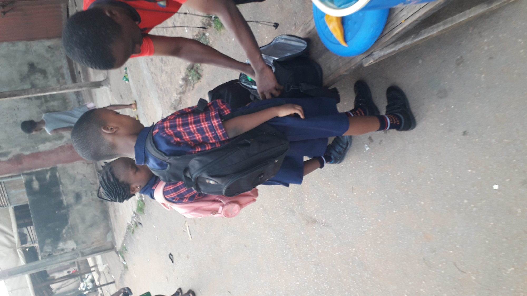 In Onitsha, Nigeria, a medium shot depicts two young children dressed in school uniforms, wearing backpacks. An adult wearing a red shirt is seen from the shoulders up, adjusting items inside a black backpack. A third child is visible in the background. A blue bucket with a yellow item inside rests on a structure above the group. The background reveals a cityscape with buildings and concrete. The surface under the children appears to be a paved area.