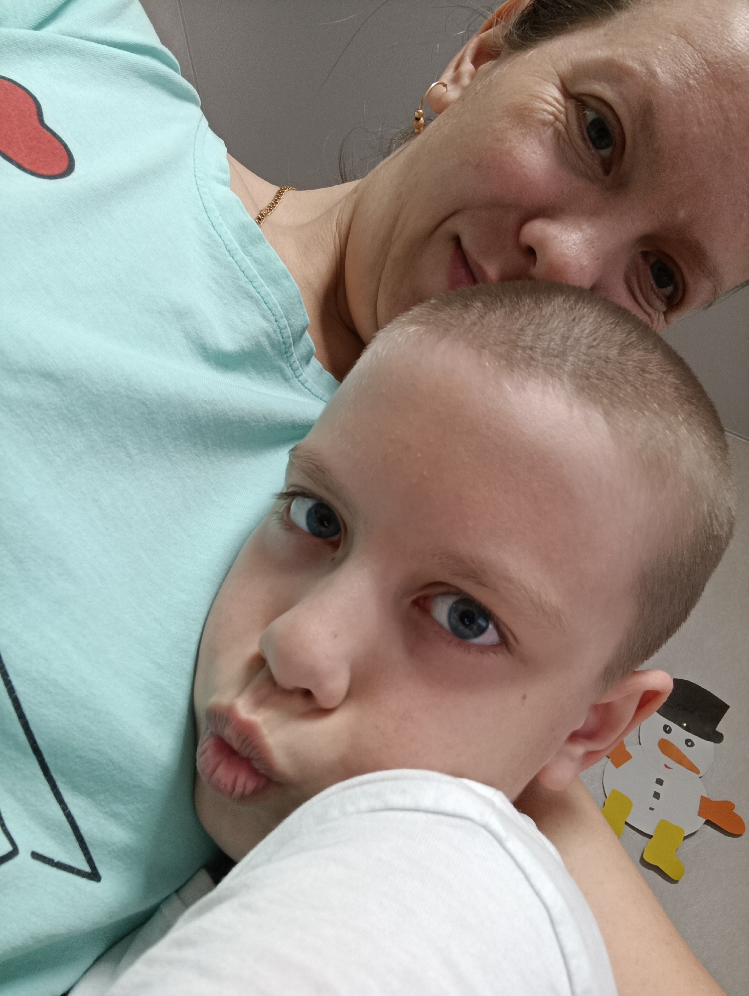 A close-up, high-angle shot depicts a fair-skinned woman with light brown hair and a gold chain around her neck, and a fair-skinned boy with close-cropped light brown hair and blue eyes. The boy rests his head against the woman, who wears a light blue T-shirt with a red and black design. The boy purses his lips toward the camera. A snowman sticker is visible on the wall behind the boy. The location is unspecified but possibly Chaikovsky, Russia.
