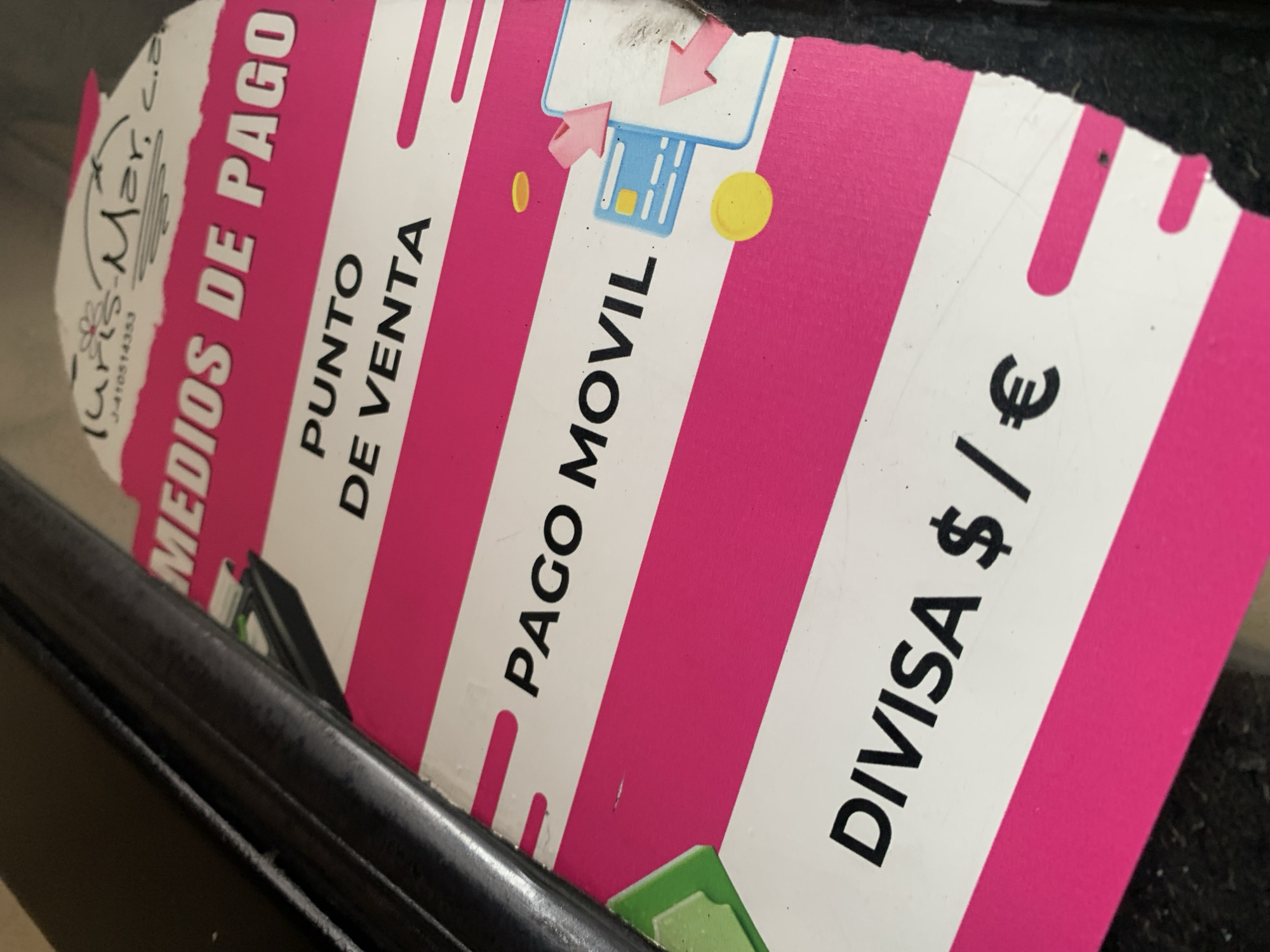 The image shows a laminated sign, likely affixed to a point-of-sale device or payment terminal. The sign is printed with alternating pink and white horizontal stripes. Text on the sign, in Spanish, includes: "Medios de Pago" (Means of Payment), "Punto de Venta" (Point of Sale), "Pago Movil" (Mobile Payment), and "Divisa $ / €" (Currency $ / €). A graphic representing a credit card with arrows indicates card payment acceptance. In the upper left corner, there is an indication to Juris-Mar, C.A. and its tax number J-410514353. The photo was taken in Tacarigua, Venezuela. The sign appears to be partially obscured by a dark, metallic object or surface.