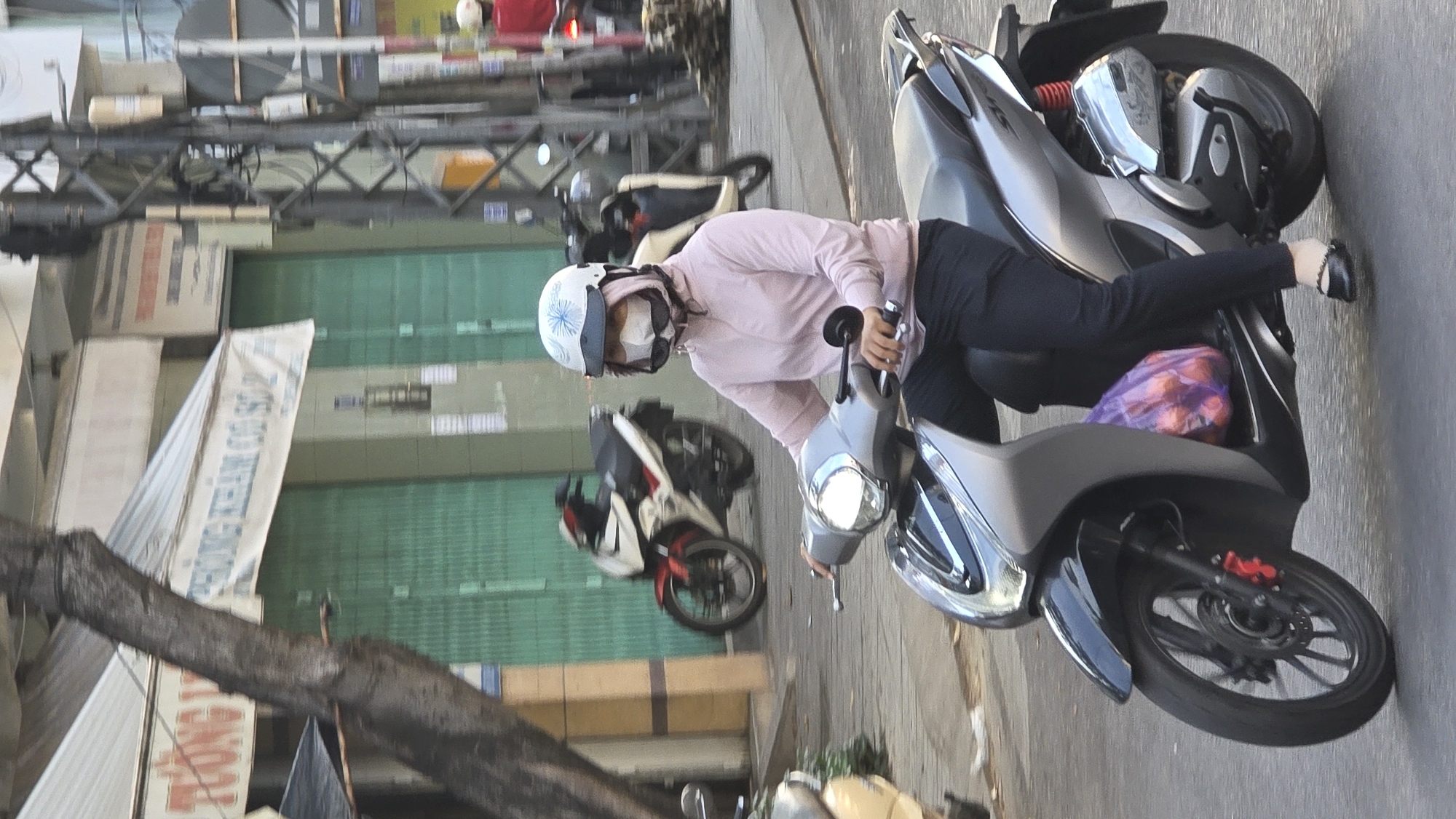 A woman rides a scooter on a city street in Nha Trang, Vietnam. The woman wears a light pink hooded shirt, dark pants, and a white helmet with a blue design, sunglasses and a white face mask. She rides a grey scooter with a bag of fruit tucked behind her leg. The street includes a tree branch in the foreground, buildings with green metal roll-up doors, and other parked scooters. Traffic is visible further down the street.