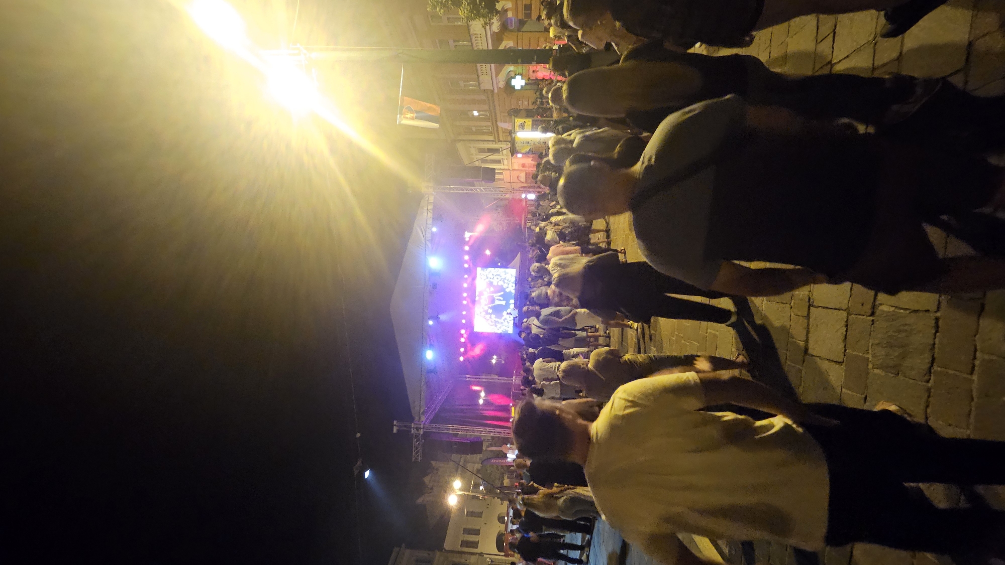 The image depicts an outdoor concert event at night in Prešov, Slovakia. A large crowd of people are gathered, facing a stage set up in a city square. The stage features a large LED screen displaying an image, and is illuminated by vibrant pink and purple stage lighting. The surrounding buildings appear to be historic, with the architecture characteristic of a European city center. A bright streetlamp illuminates the scene from the right side of the image, casting a strong glare. The ground is paved with cobblestones.