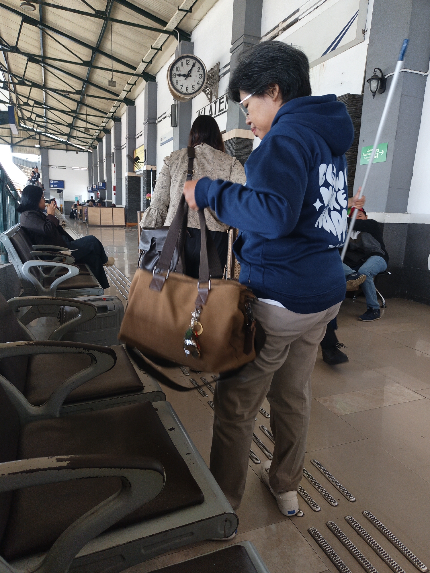 A medium shot shows a woman in a blue hoodie and tan pants walking away from the camera at a train station in Klaten, Indonesia. She carries a large tan duffel bag over her shoulder. In the background, a clock on the wall reads approximately 10:08. Other passengers are visible seated on benches and standing. The environment includes a covered platform with a metal support structure overhead.