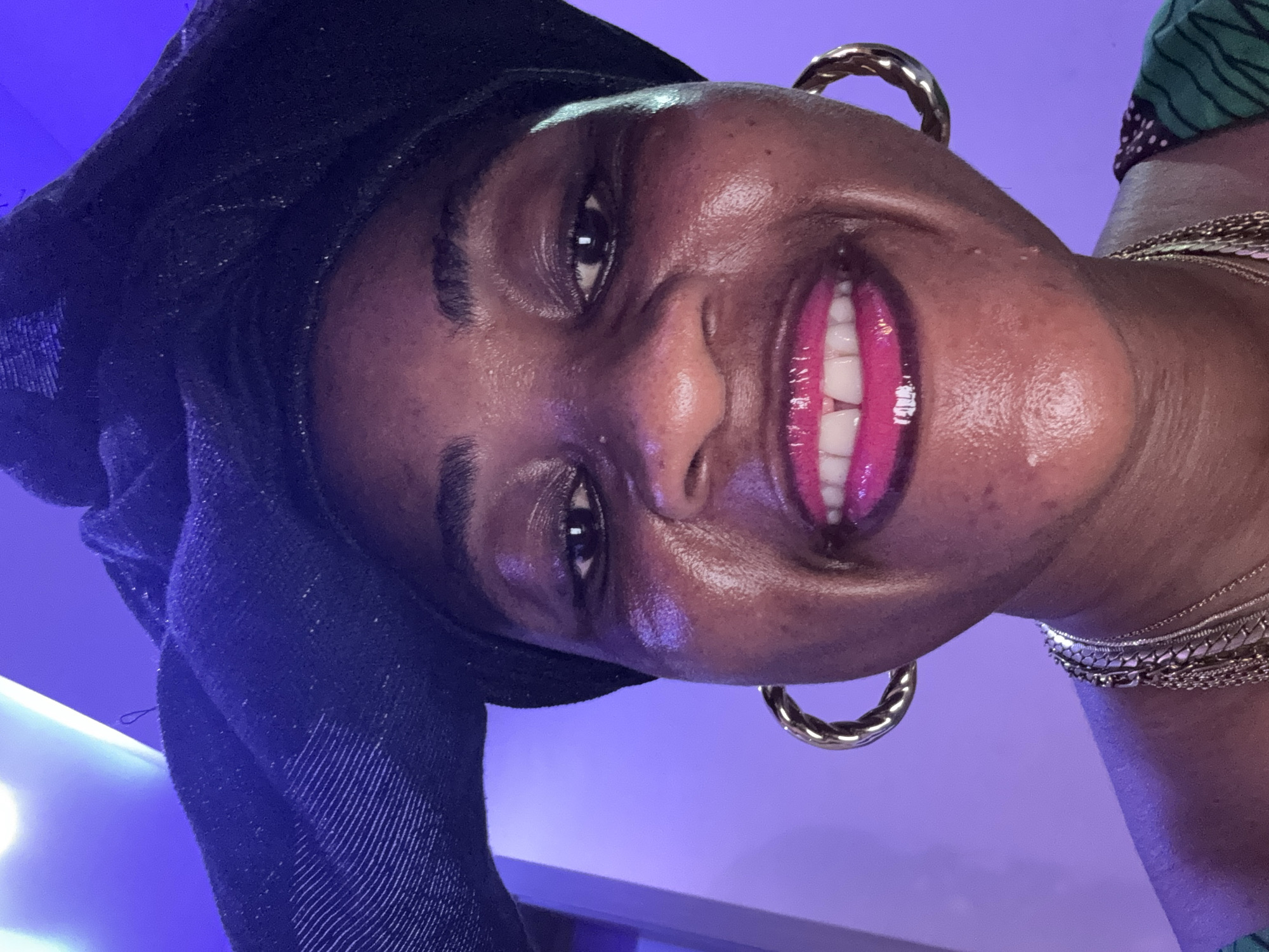 A close-up, front-facing selfie shows a Black woman in a black headwrap and large gold hoop earrings. She is wearing multiple layered gold necklaces and a green garment with a dark pattern. Her lips are painted a bright fuchsia. The background is a solid purple wall with a bright blue light source visible in the upper left corner. The image appears to have been taken indoors, possibly in a home or event space. There is no visible indication of the specific location within Ajara, Nigeria.