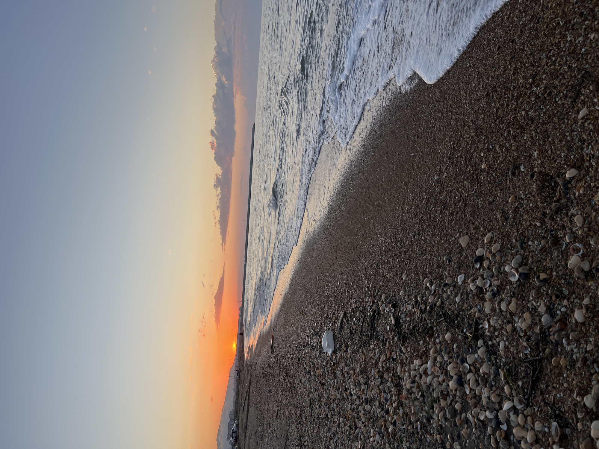 The image depicts a beach scene in Kaspiysk, Russia, during a sunset. The sky transitions from light blue to orange near the horizon where the sun is setting. Clouds are present along the horizon. Waves are visible in the sea, approaching the shore. The beach consists of sand and pebbles, with a high concentration of seashells towards the lower portion of the image. A discarded plastic container is visible on the beach. The image is rotated.