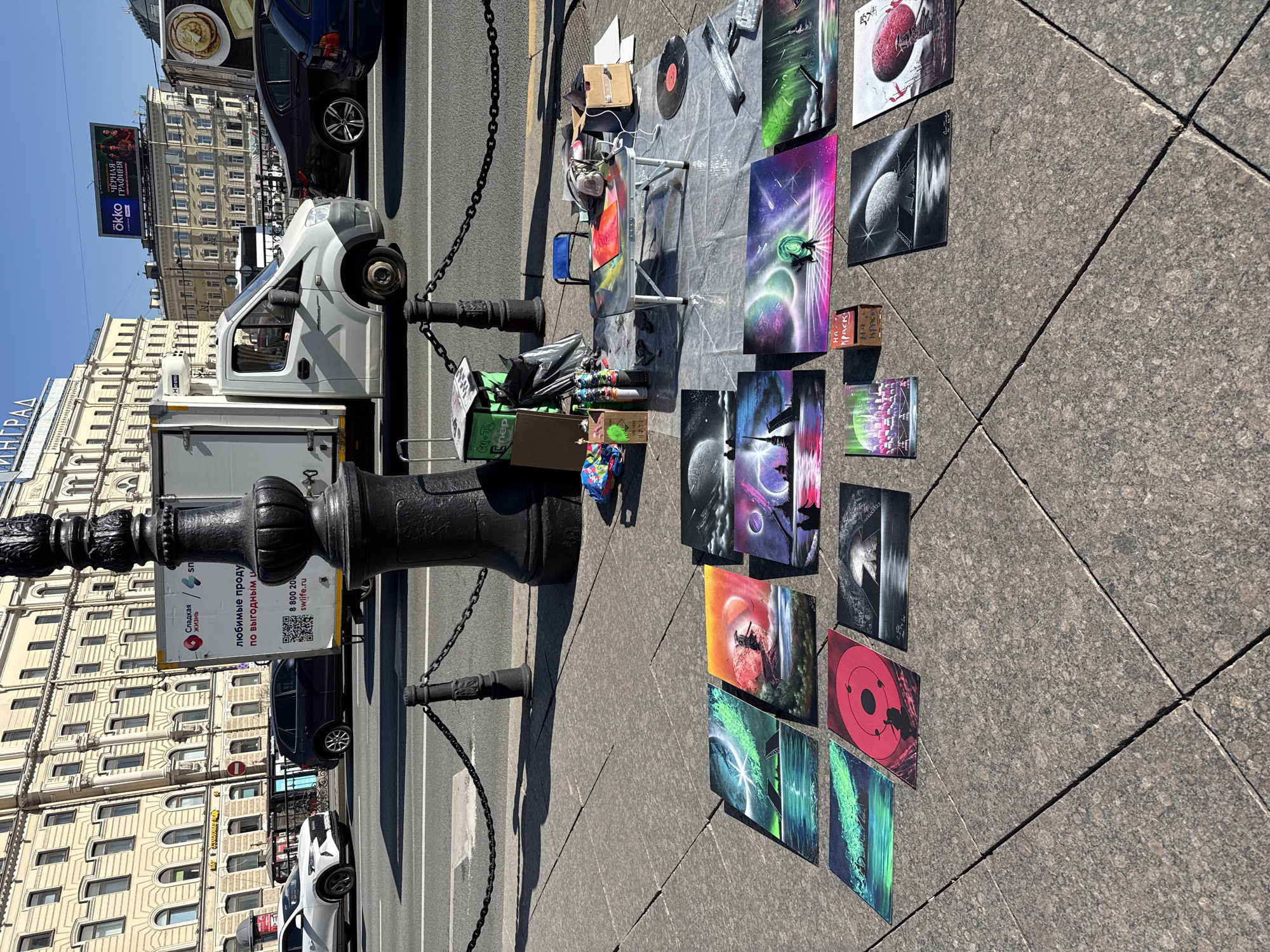 The image shows an outdoor street scene in Saint Petersburg, Russia. A vendor has set up an art display on the sidewalk consisting of a variety of paintings and spray-painted artwork laid out on the pavement. The paintings feature various subjects including cosmic scenes, planets, and abstract designs. Alongside the artwork, there is a table with art supplies, cans, brushes, and tools. A vinyl record can be seen on the table, which indicates the work setup.
A black, ornate metal bollard with chains is situated near the artwork display, separating the sidewalk from the street. Vehicles, including a white delivery van and passenger cars, are visible on the adjacent street. Buildings are visible in the background, including a structure with the word "LENINGRAD" on it, a billboard with "Ökko" and "Черная графиня" visible on it. A smaller sign with a QR code can be seen attached to the bollard. The sidewalk has a patterned, tiled surface.